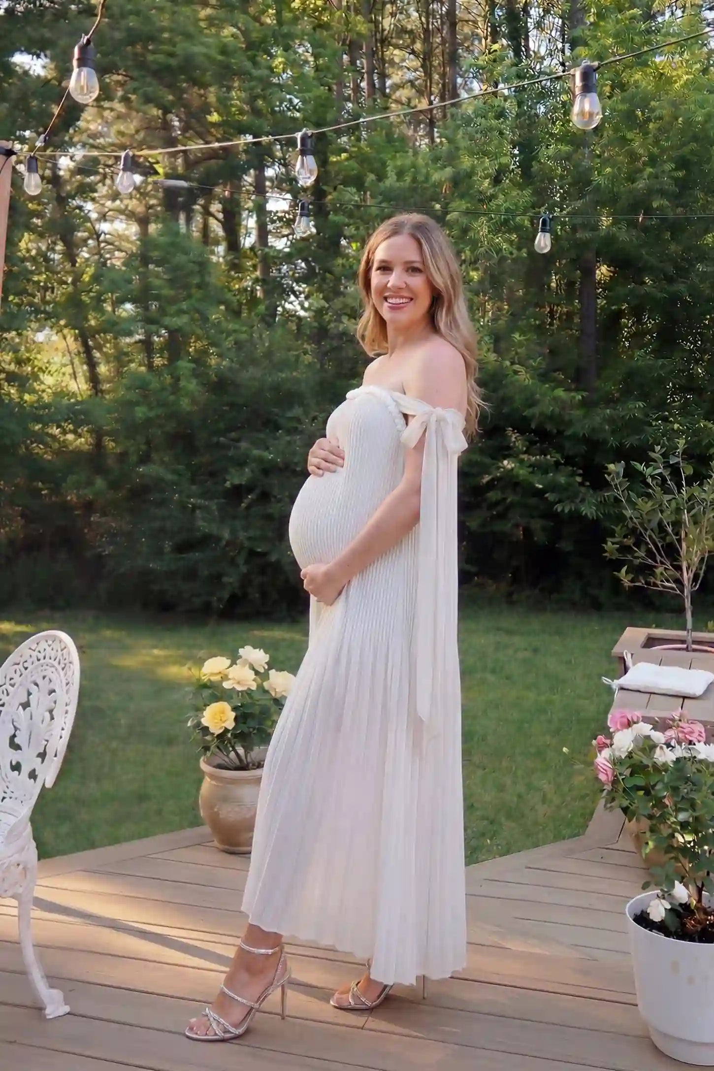 Pleated Braided Cold-Shoulder Maxi Maternity Gown Pleated Braided Cold-Shoulder Maxi Maternity Gown in White featuring soft pleated fabric on a pregnant model in an editorial lifestyle setting.