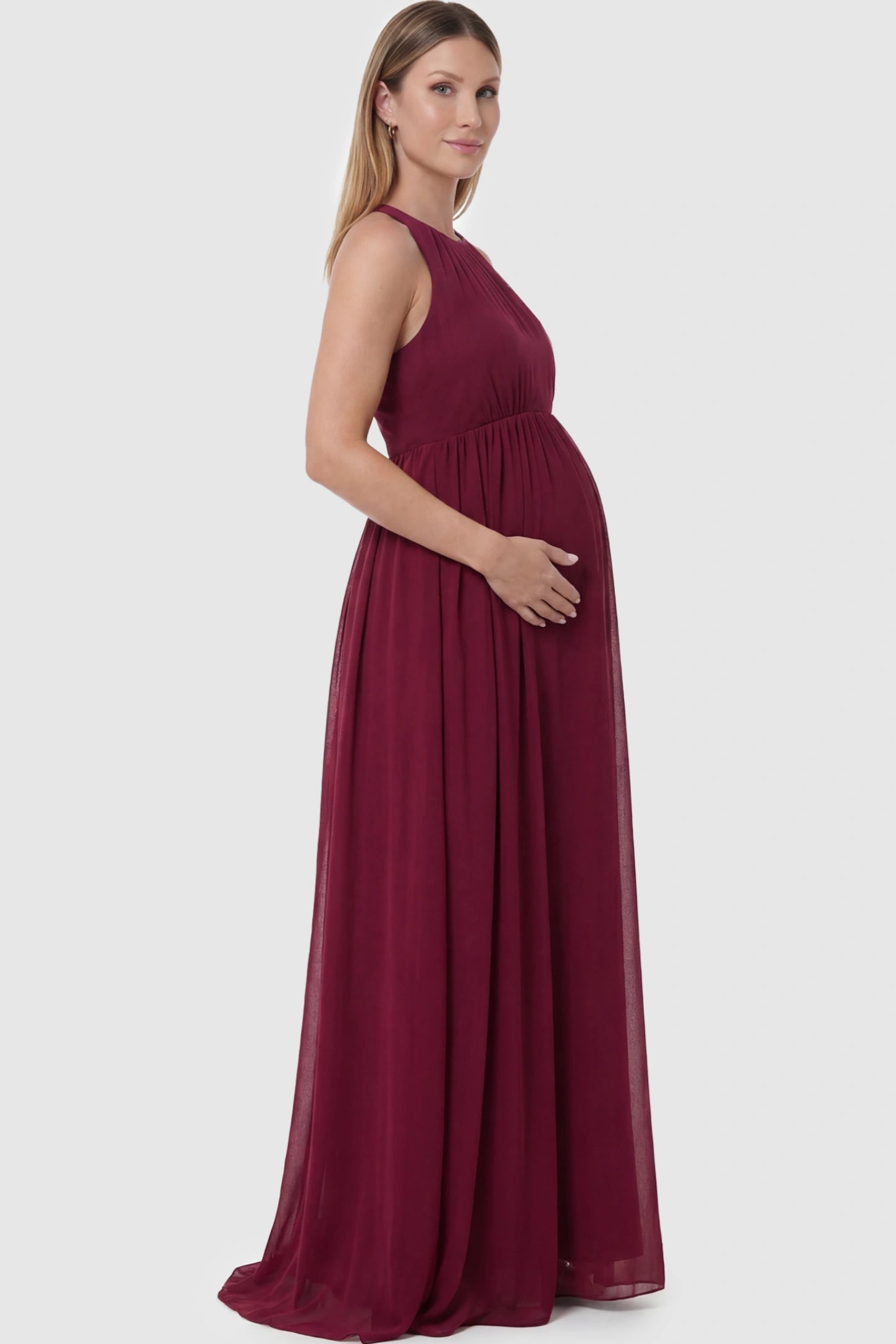 Pleated Mesh Sleeveless Maternity Photoshoot Dress Pleated Mesh Sleeveless Maternity Maxi Dress in burgundy featuring an empire waist on a pregnant model showing side.