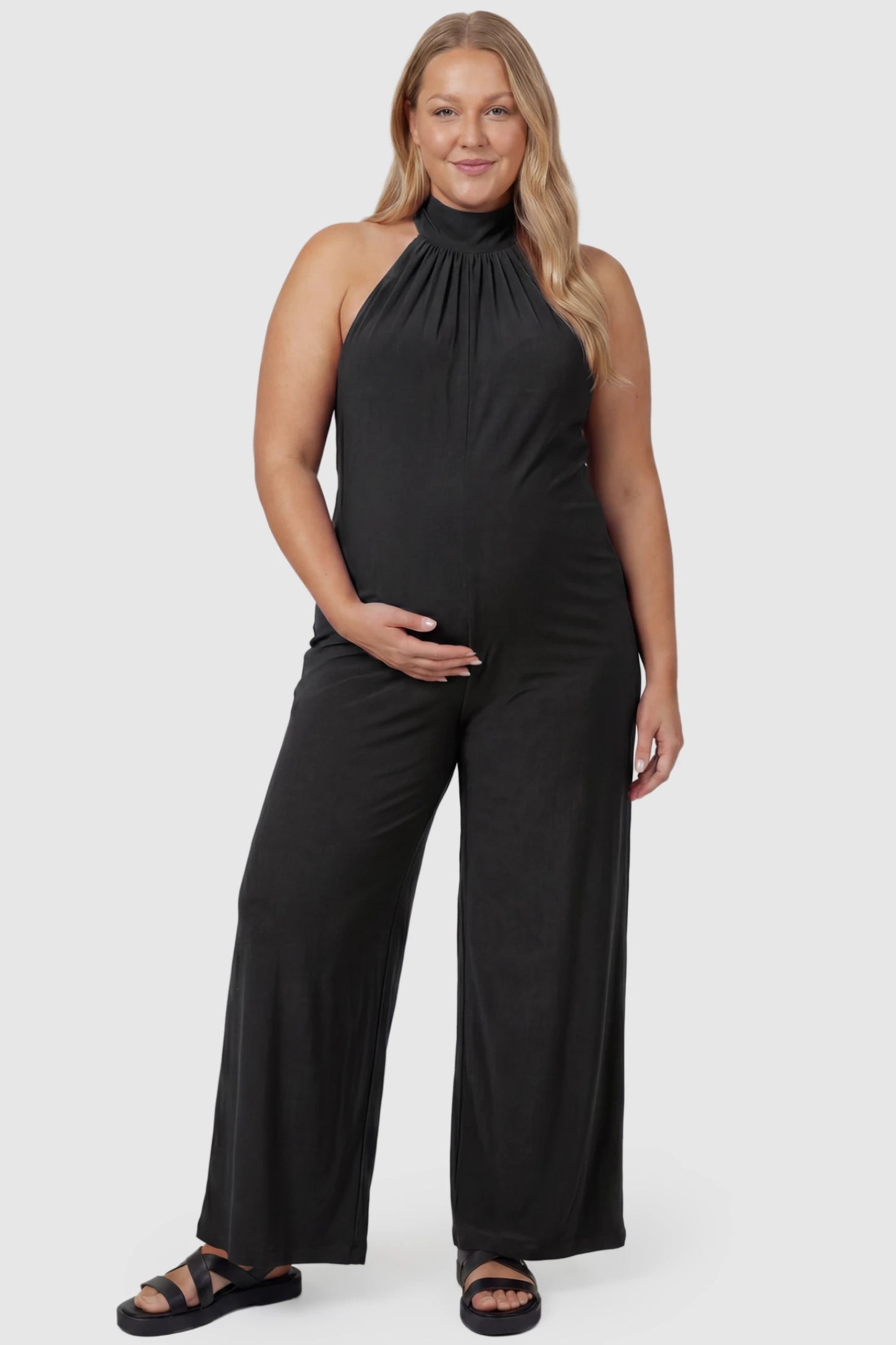 Pocketed Mock Neck Sleeveless Maternity Jumpsuit Pocketed Mock Neck Sleeveless Maternity Jumpsuit in black featuring a high gathered neckline, side pockets shown on a plus size pregnant model, front view.