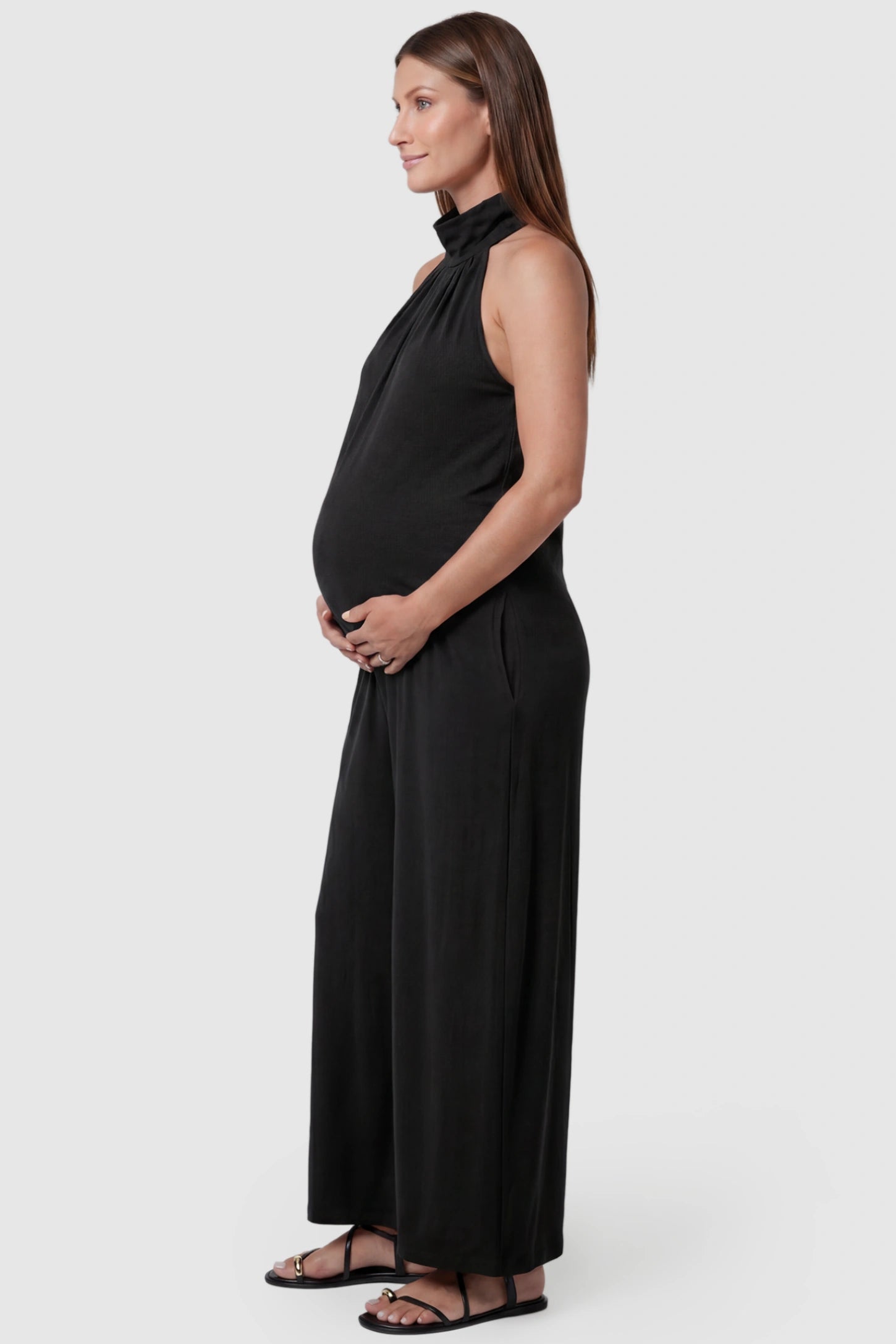 Pocketed Mock Neck Sleeveless Maternity Jumpsuit Pocketed Mock Neck Sleeveless Maternity Jumpsuit in black featuring a high gathered neckline, side pockets shown on a pregnant model, side view.