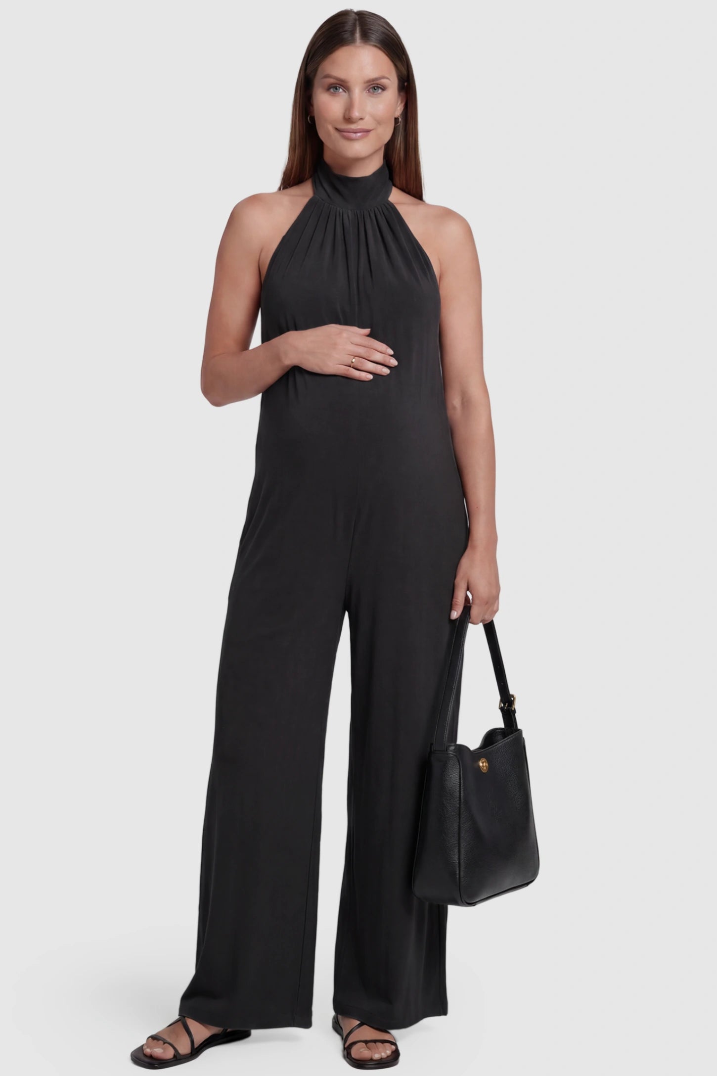Pocketed Mock Neck Sleeveless Maternity Jumpsuit Pocketed Mock Neck Sleeveless Maternity Jumpsuit in black featuring a high gathered neckline, side pockets shown on a pregnant model holding a black shoulder bag, front view.