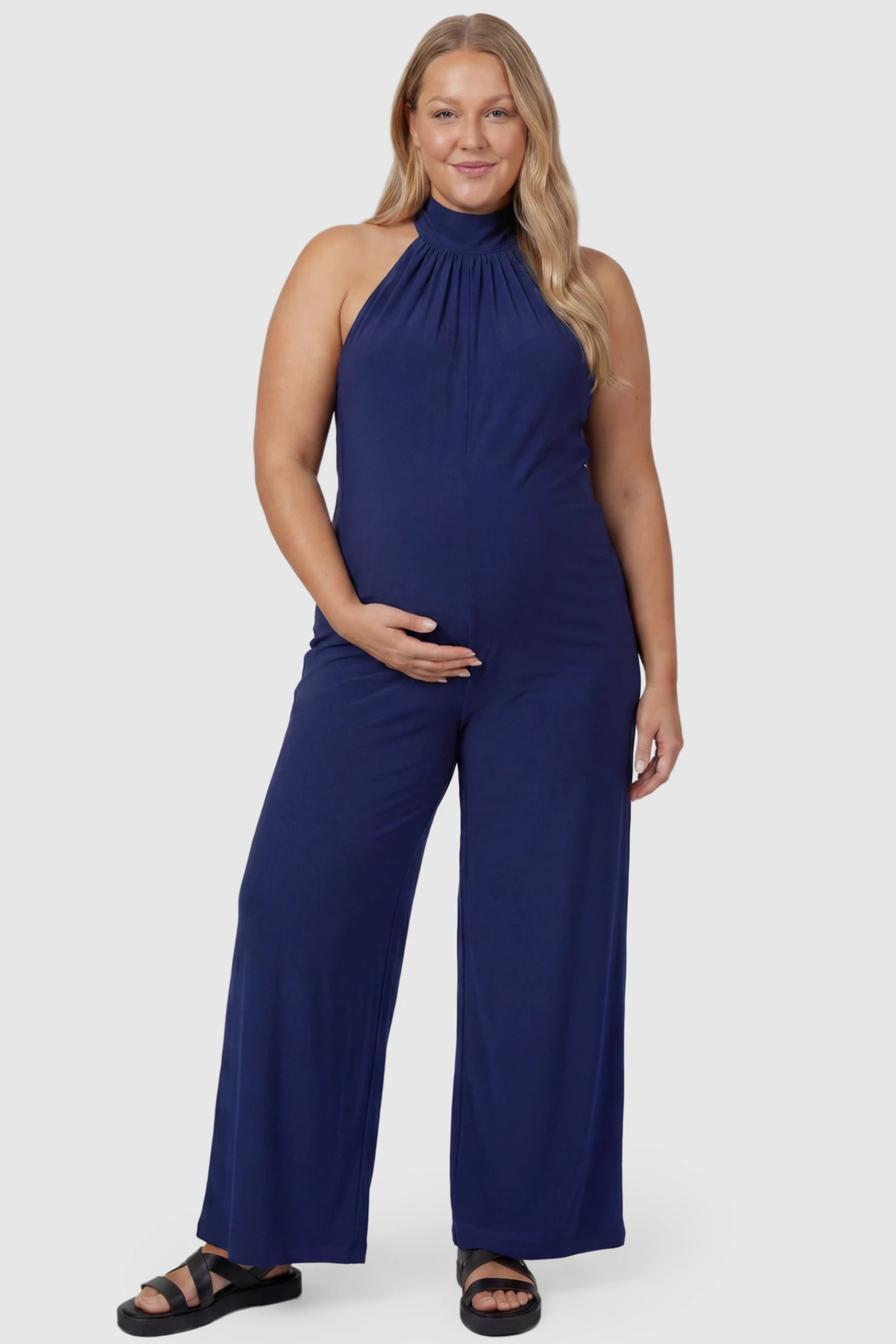 Pocketed Mock Neck Sleeveless Maternity Jumpsuit Pocketed Mock Neck Sleeveless Maternity Jumpsuit in blue featuring a high gathered neckline, side pockets shown on a plus size pregnant model, front view.