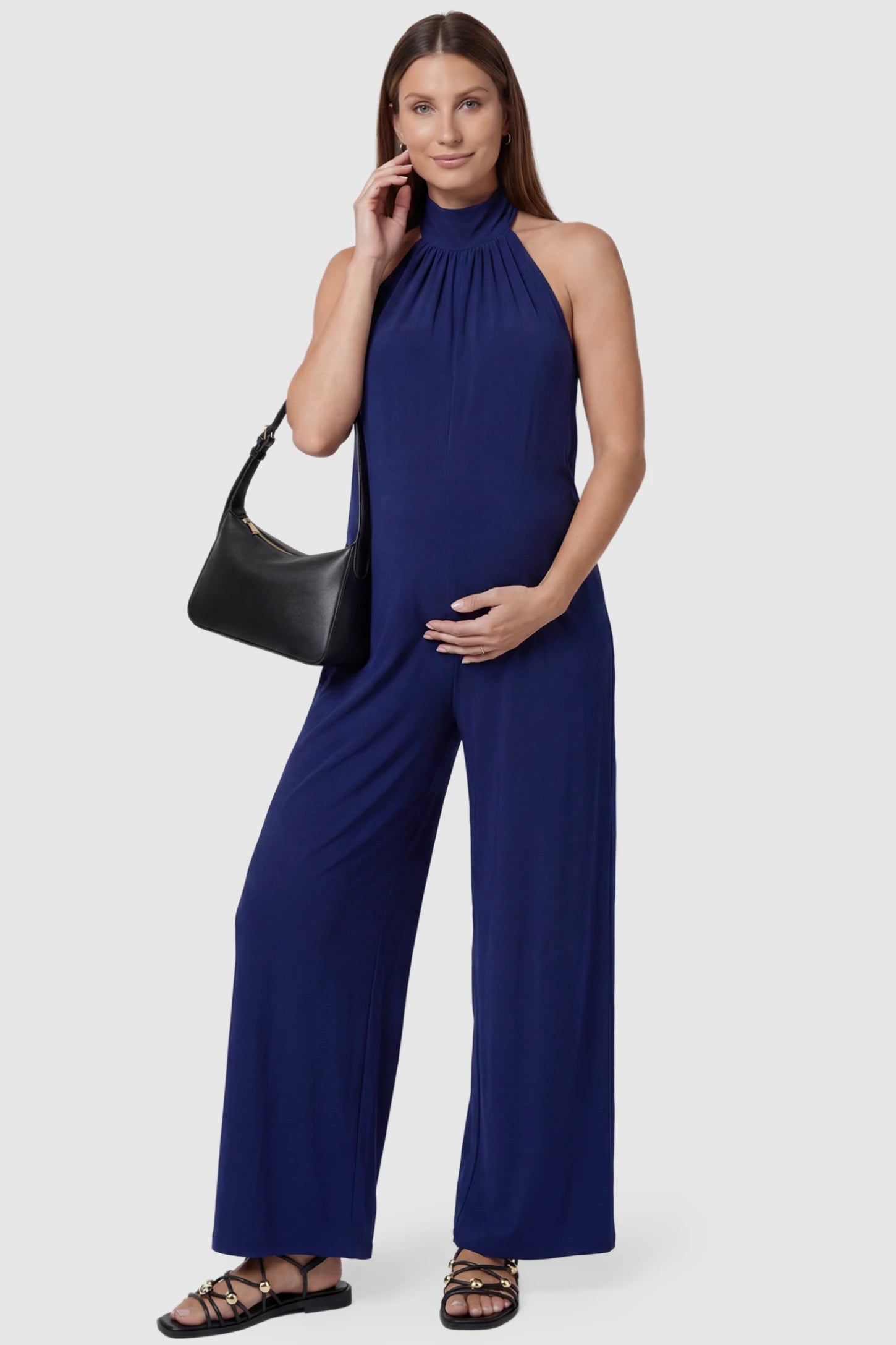 Pocketed Mock Neck Sleeveless Maternity Jumpsuit Pocketed Mock Neck Sleeveless Maternity Jumpsuit in blue featuring a high gathered neckline, side pockets shown on a pregnant model holding a black shoulder bag, front view.