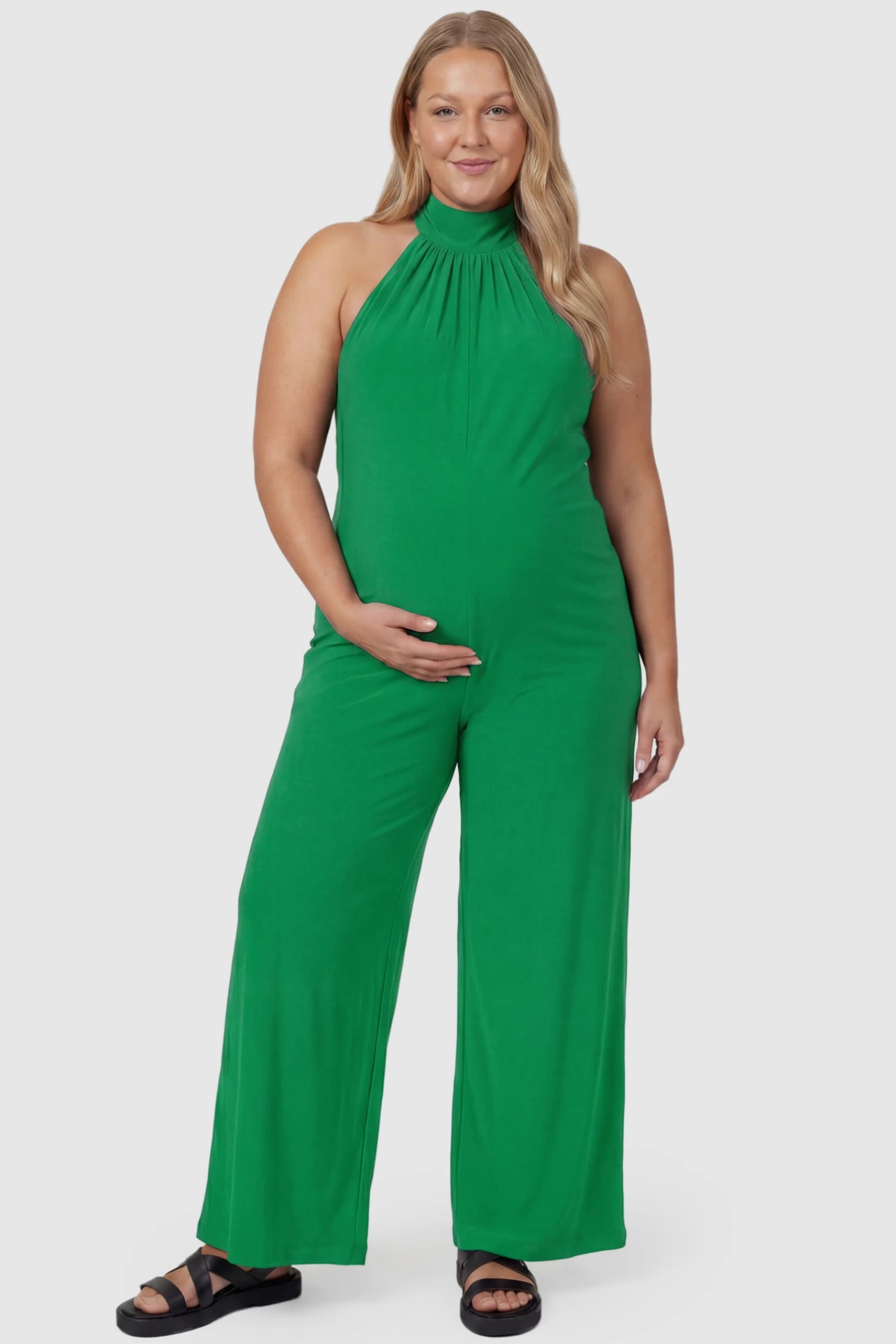 Pocketed Mock Neck Sleeveless Maternity Jumpsuit Pocketed Mock Neck Sleeveless Maternity Jumpsuit in green featuring a high gathered neckline, side pockets shown on a plus size pregnant model, front view.