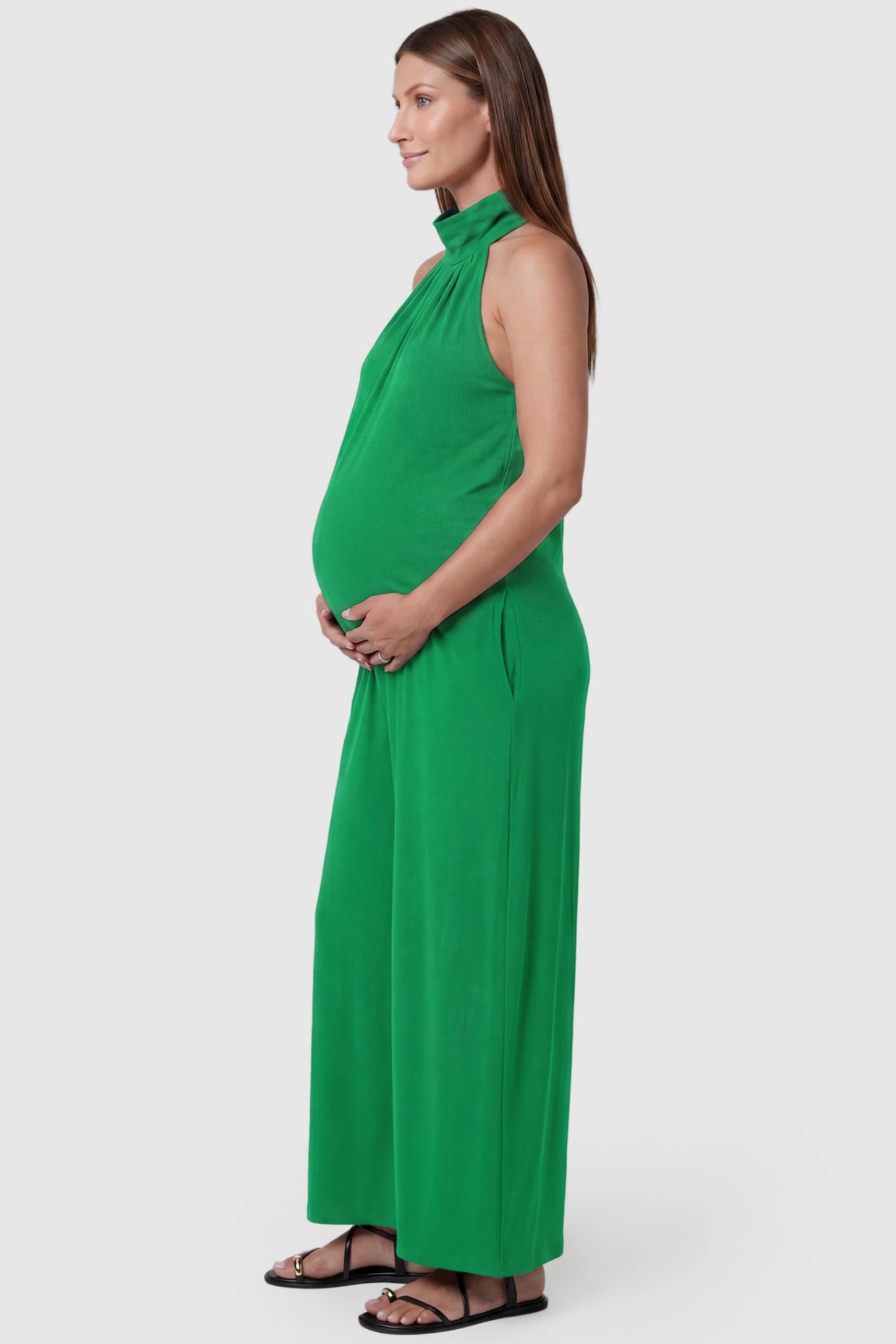 Pocketed Mock Neck Sleeveless Maternity Jumpsuit Pocketed Mock Neck Sleeveless Maternity Jumpsuit in green featuring a high gathered neckline, side pockets shown on a pregnant model, side view.