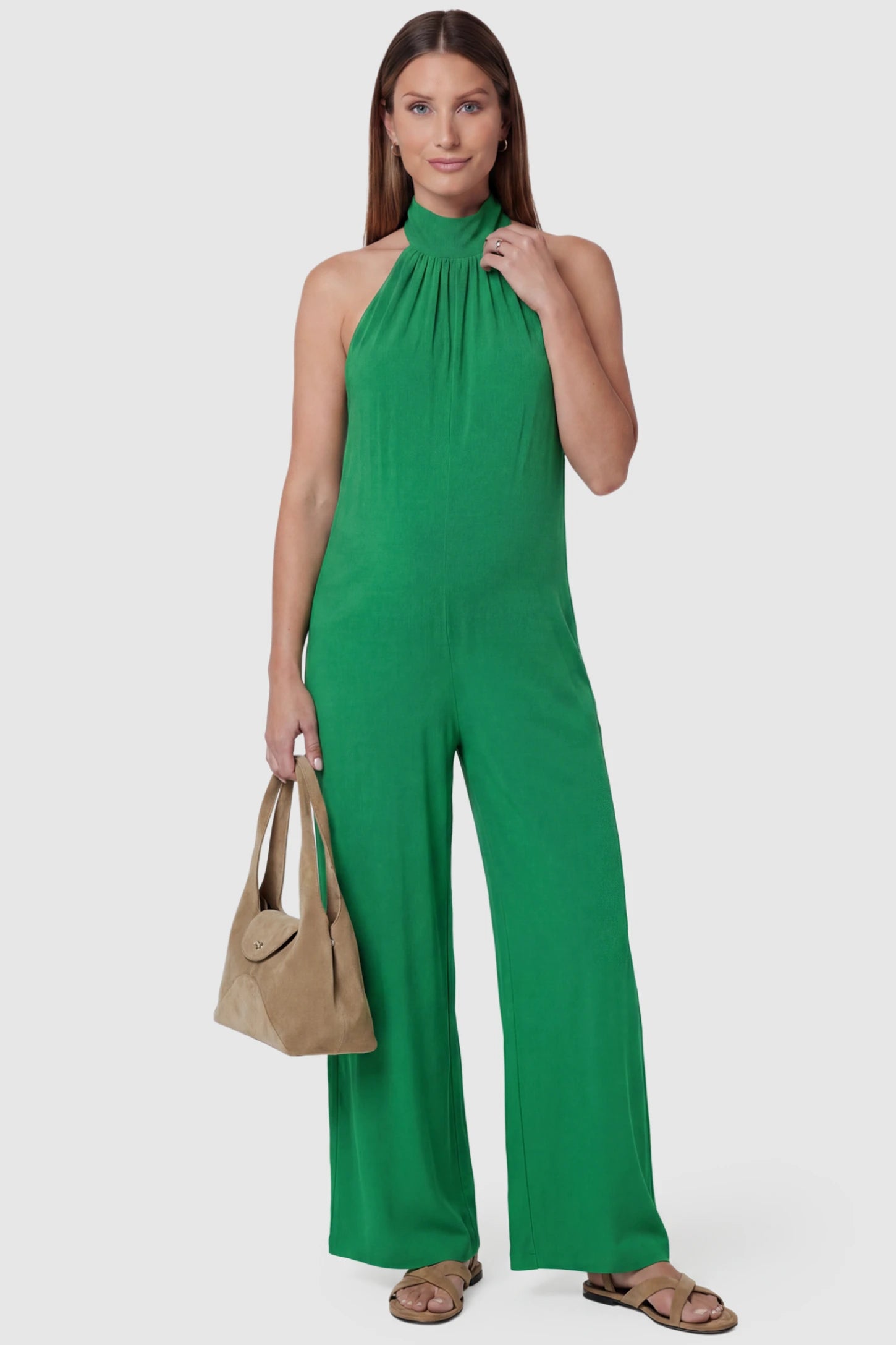 Pocketed Mock Neck Sleeveless Maternity Jumpsuit Pocketed Mock Neck Sleeveless Maternity Jumpsuit in green featuring a high gathered neckline, side pockets shown on a pregnant model holding a bag, front view.