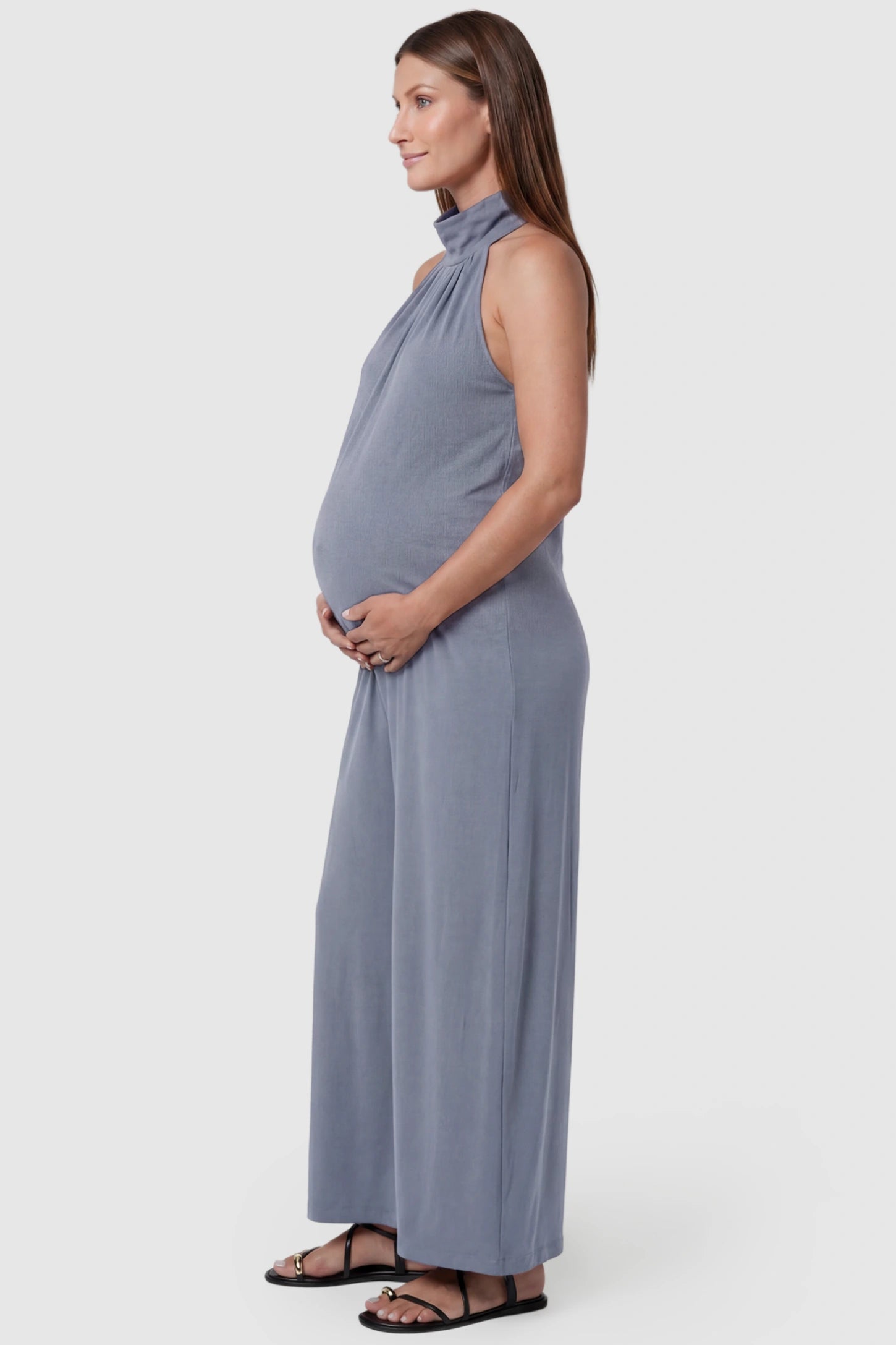 Pocketed Mock Neck Sleeveless Maternity Jumpsuit Pocketed Mock Neck Sleeveless Maternity Jumpsuit in light blue featuring a high gathered neckline, side pockets shown on a pregnant model, side view.