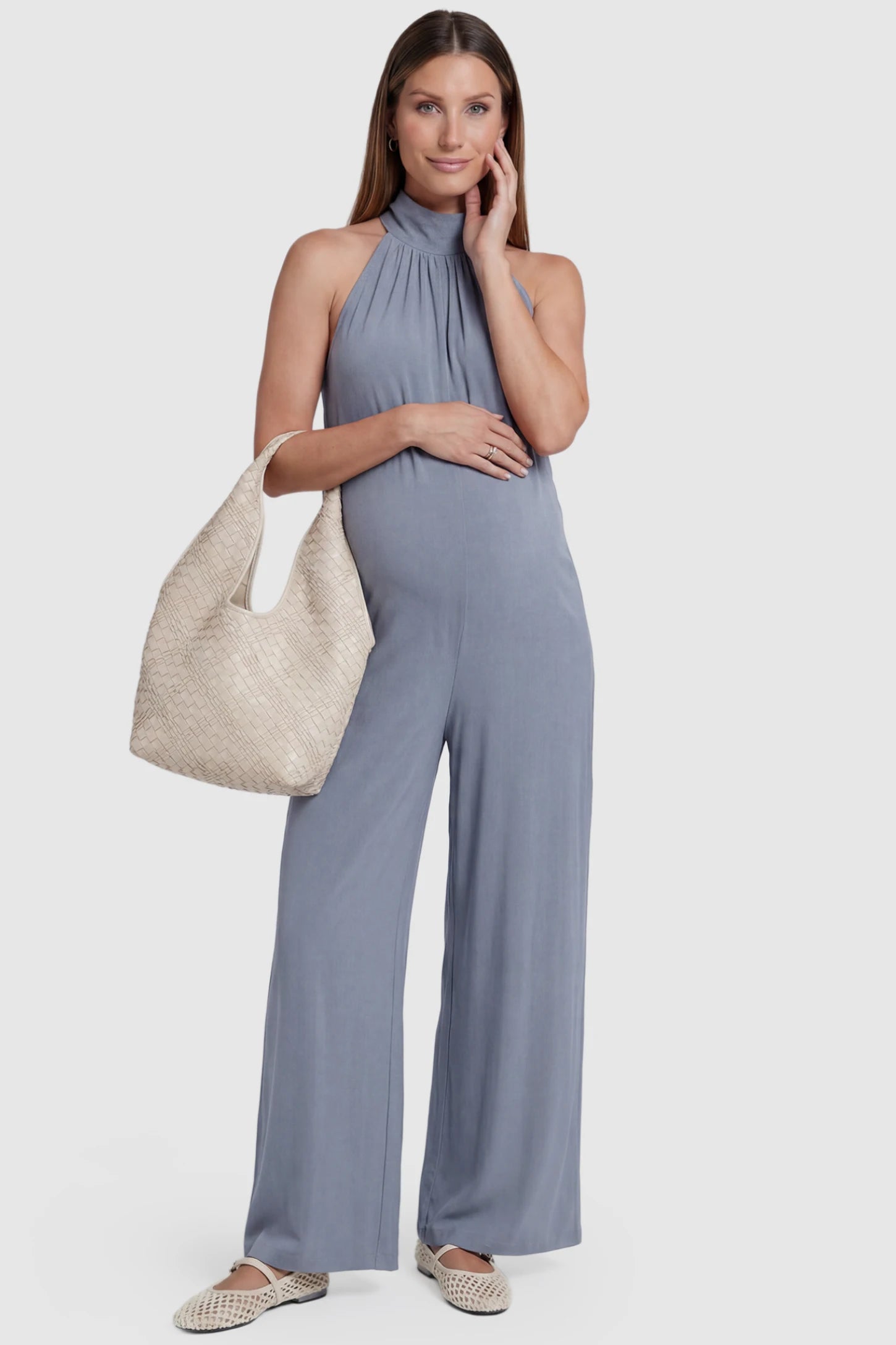 Pocketed Mock Neck Sleeveless Maternity Jumpsuit Pocketed Mock Neck Sleeveless Maternity Jumpsuit in light blue featuring a high gathered neckline, side pockets shown on a pregnant model holding a beige bag, front view.