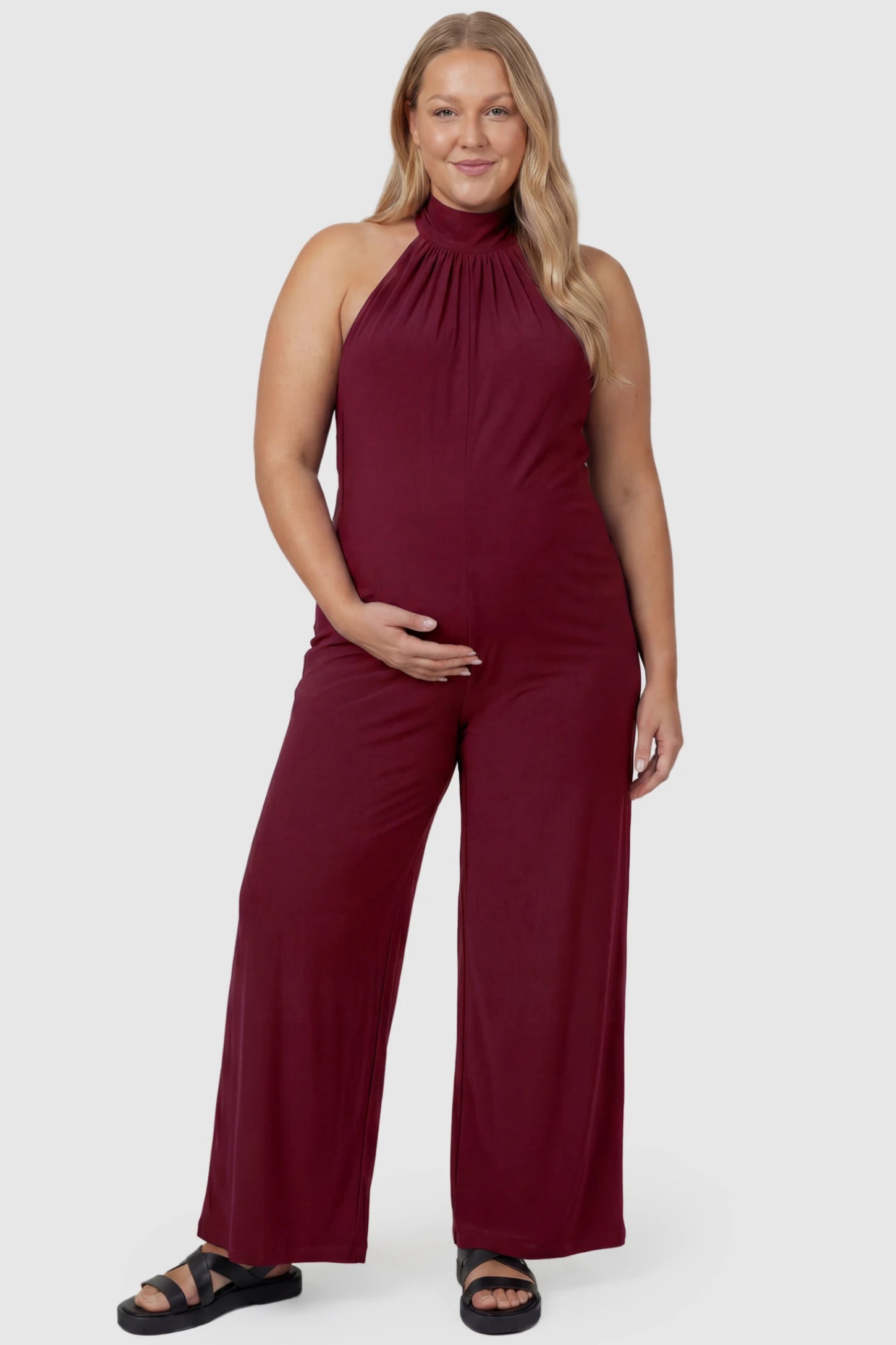 Pocketed Mock Neck Sleeveless Maternity Jumpsuit Pocketed Mock Neck Sleeveless Maternity Jumpsuit in rose red featuring a high gathered neckline, side pockets shown on a plus size pregnant model, front view.