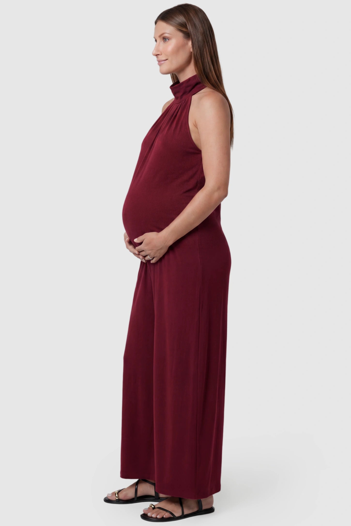 Pocketed Mock Neck Sleeveless Maternity Jumpsuit Pocketed Mock Neck Sleeveless Maternity Jumpsuit in rose red featuring a high gathered neckline, side pockets shown on a pregnant model, side view.