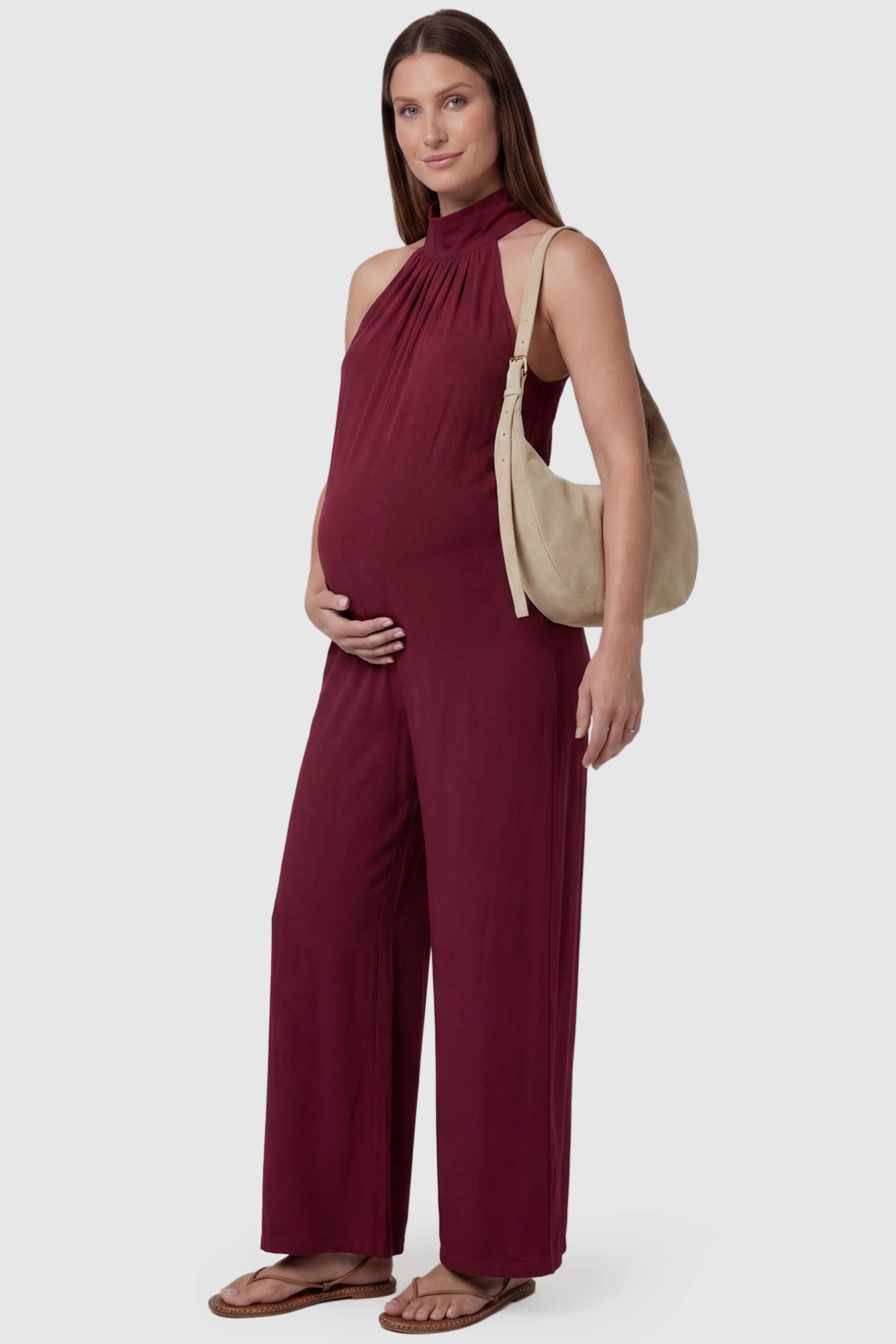 Pocketed Mock Neck Sleeveless Maternity Jumpsuit Pocketed Mock Neck Sleeveless Maternity Jumpsuit in rose red featuring a high gathered neckline, side pockets shown on a pregnant model holding a beige shoulder bag, front view.