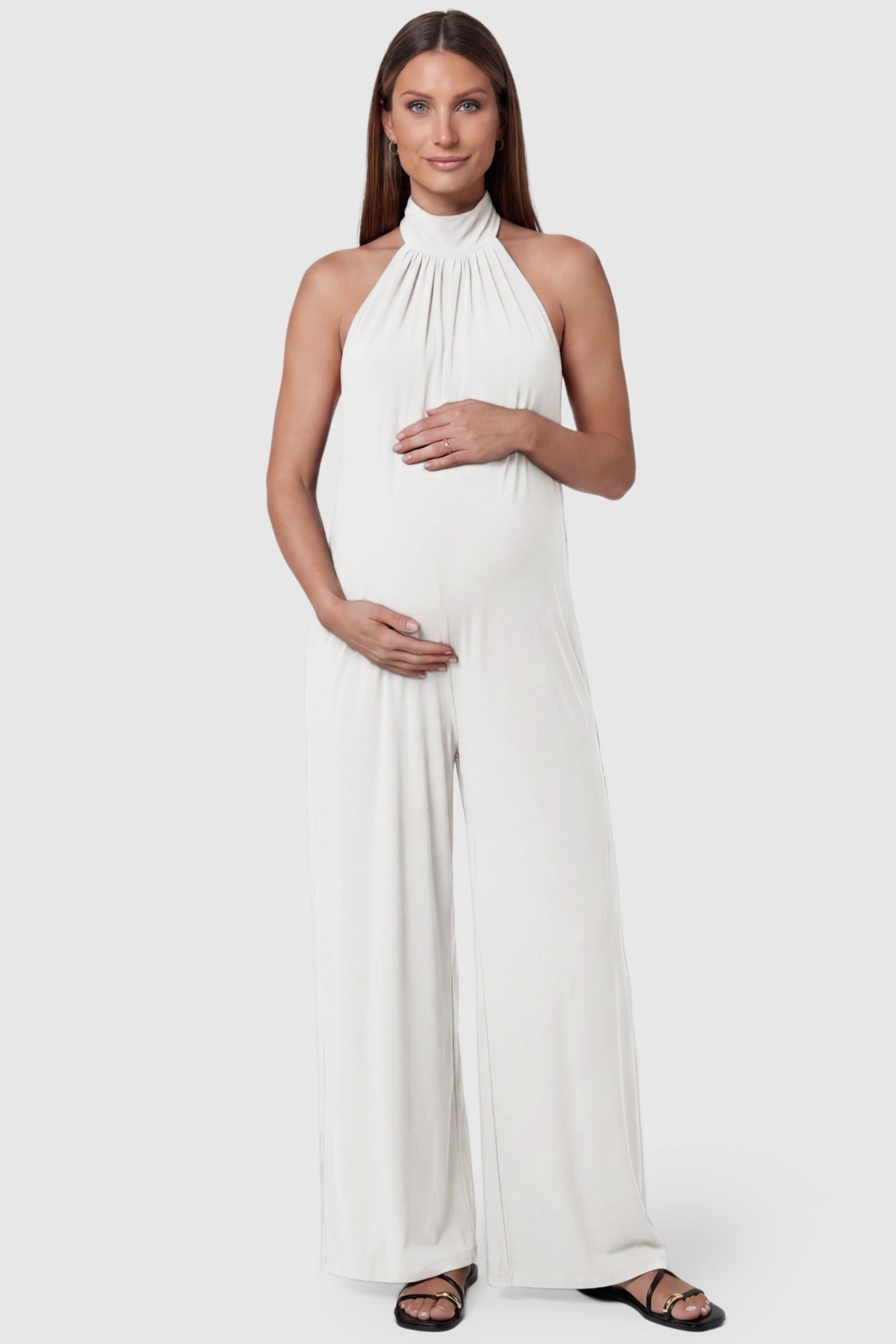 Pocketed Mock Neck Sleeveless Maternity Jumpsuit Pocketed Mock Neck Sleeveless Maternity Jumpsuit in white featuring a high gathered neckline, side pockets shown on a pregnant model, front view.
