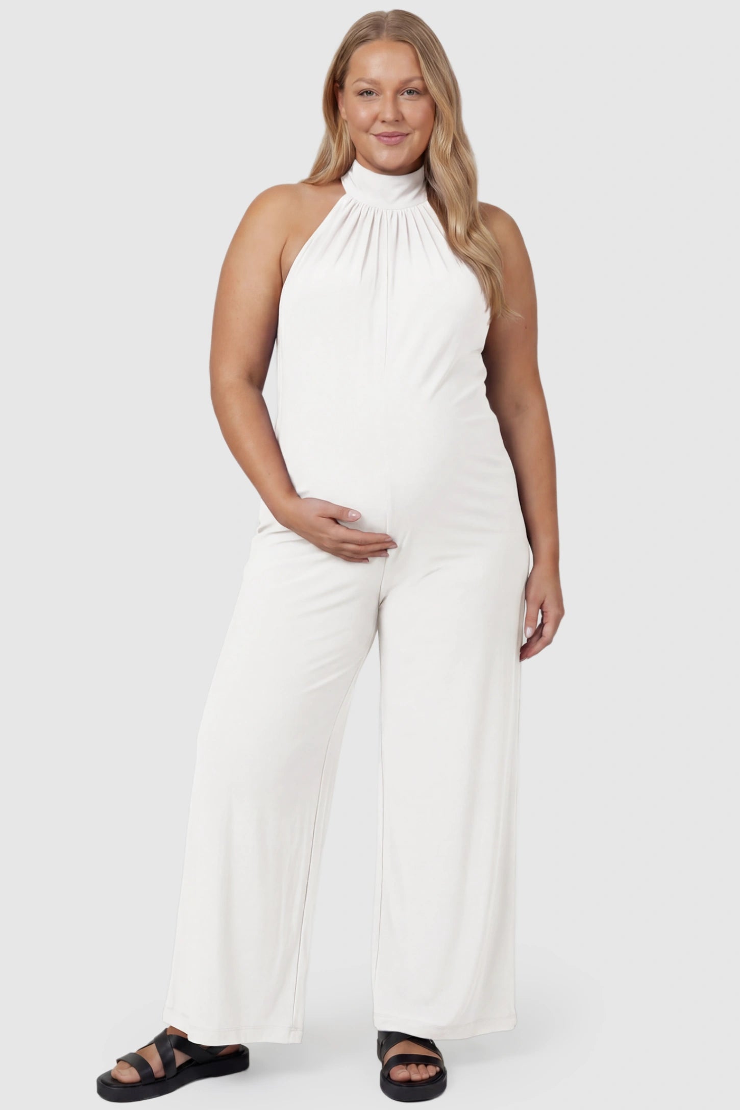 Pocketed Mock Neck Sleeveless Maternity Jumpsuit Pocketed Mock Neck Sleeveless Maternity Jumpsuit in white featuring a high gathered neckline, side pockets shown on a plus size pregnant model, front view.