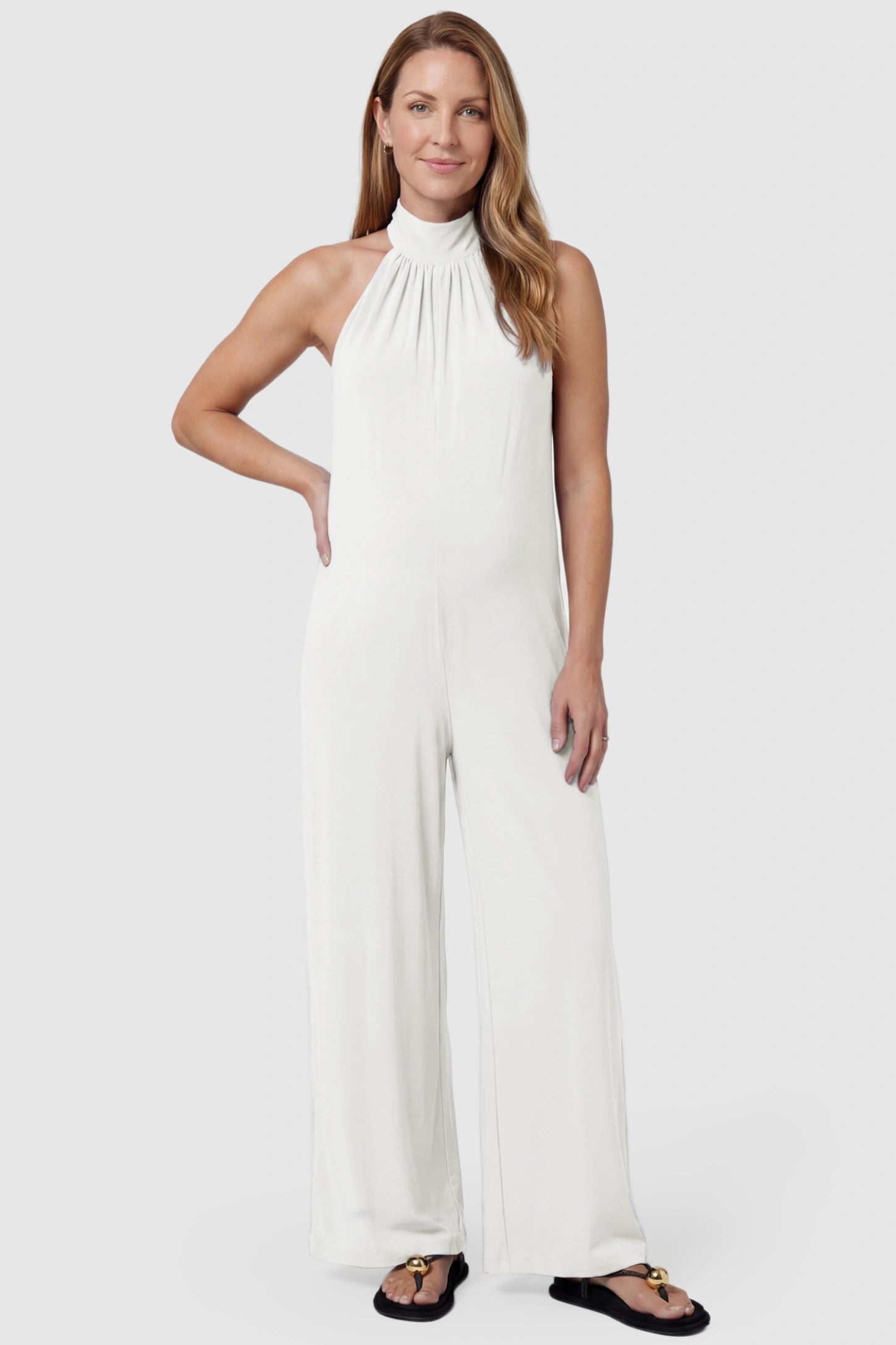 Pocketed Mock Neck Sleeveless Maternity Jumpsuit Pocketed Mock Neck Sleeveless Maternity Jumpsuit in white featuring a high gathered neckline, side pockets shown on a postpartum model, front view.
