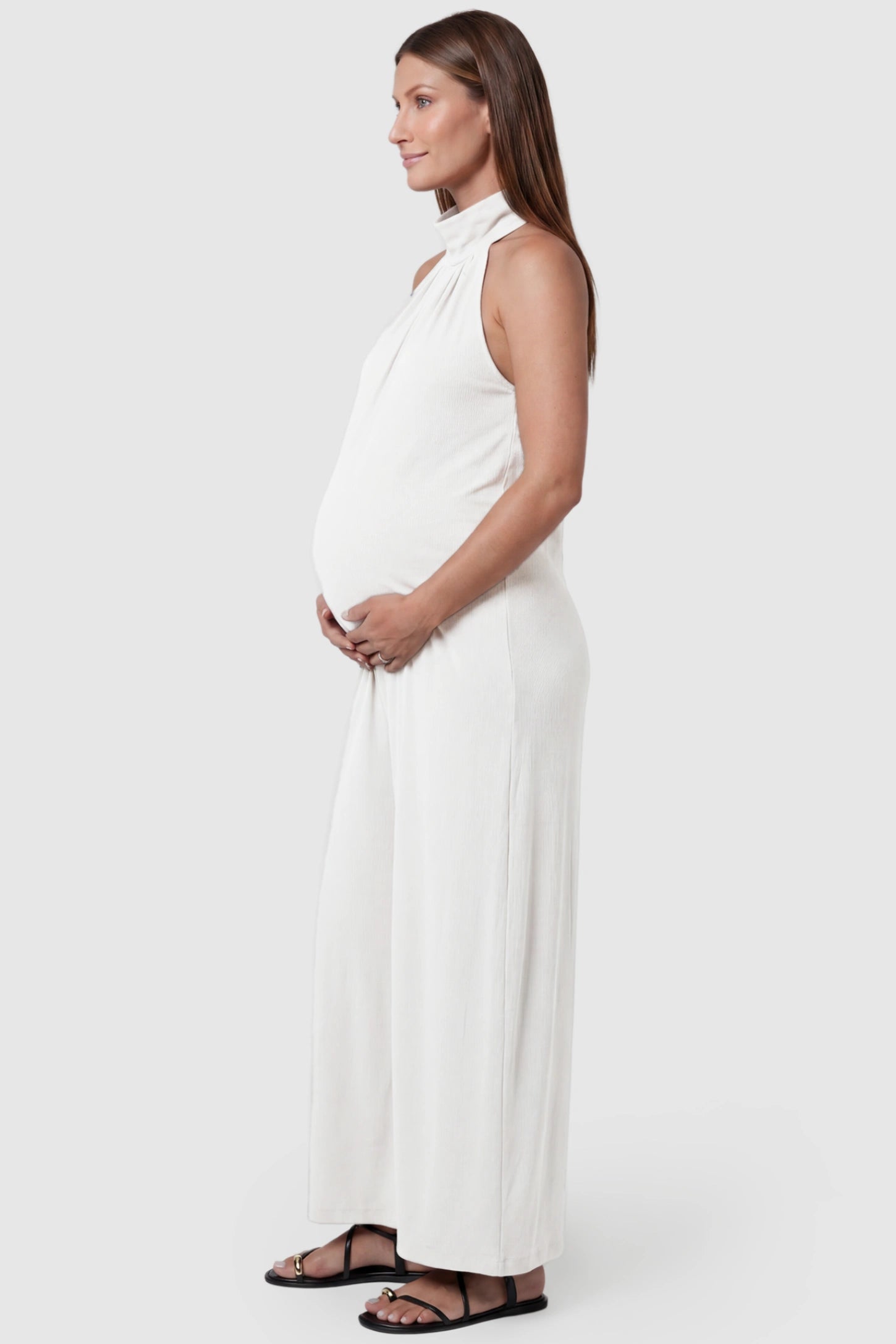Pocketed Mock Neck Sleeveless Maternity Jumpsuit Pocketed Mock Neck Sleeveless Maternity Jumpsuit in white featuring a high gathered neckline, side pockets shown on a pregnant model, side view.