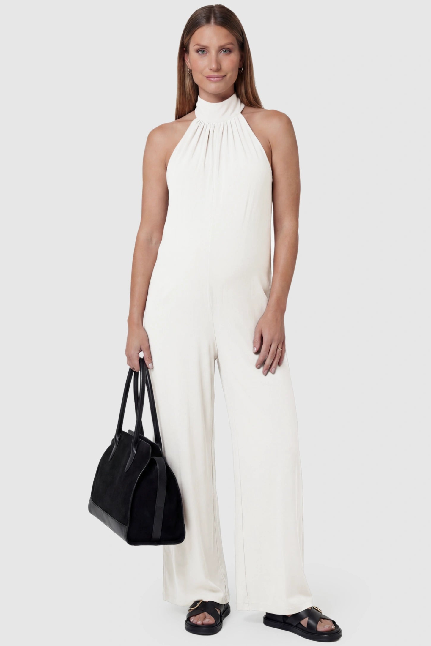 Pocketed Mock Neck Sleeveless Maternity Jumpsuit Pocketed Mock Neck Sleeveless Maternity Jumpsuit in white featuring a high gathered neckline, side pockets shown on a pregnant model holding a black bag, front view.