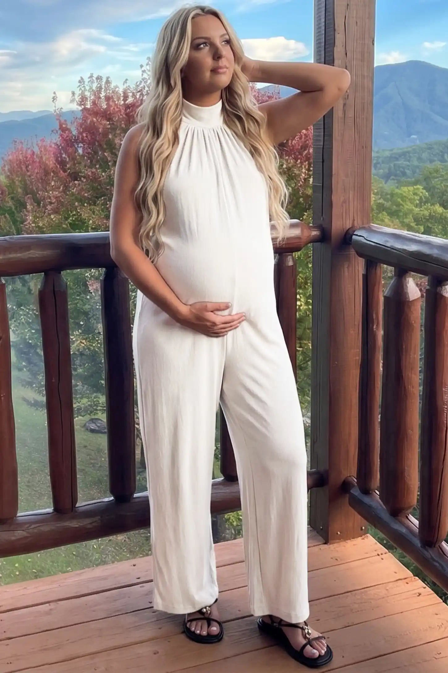 Pocketed Mock Neck Sleeveless Maternity Jumpsuit Pocketed Mock Neck Sleeveless Maternity Jumpsuit in white featuring a high gathered neckline, side pockets shown on a pregnant woman cradling her bump, side view.