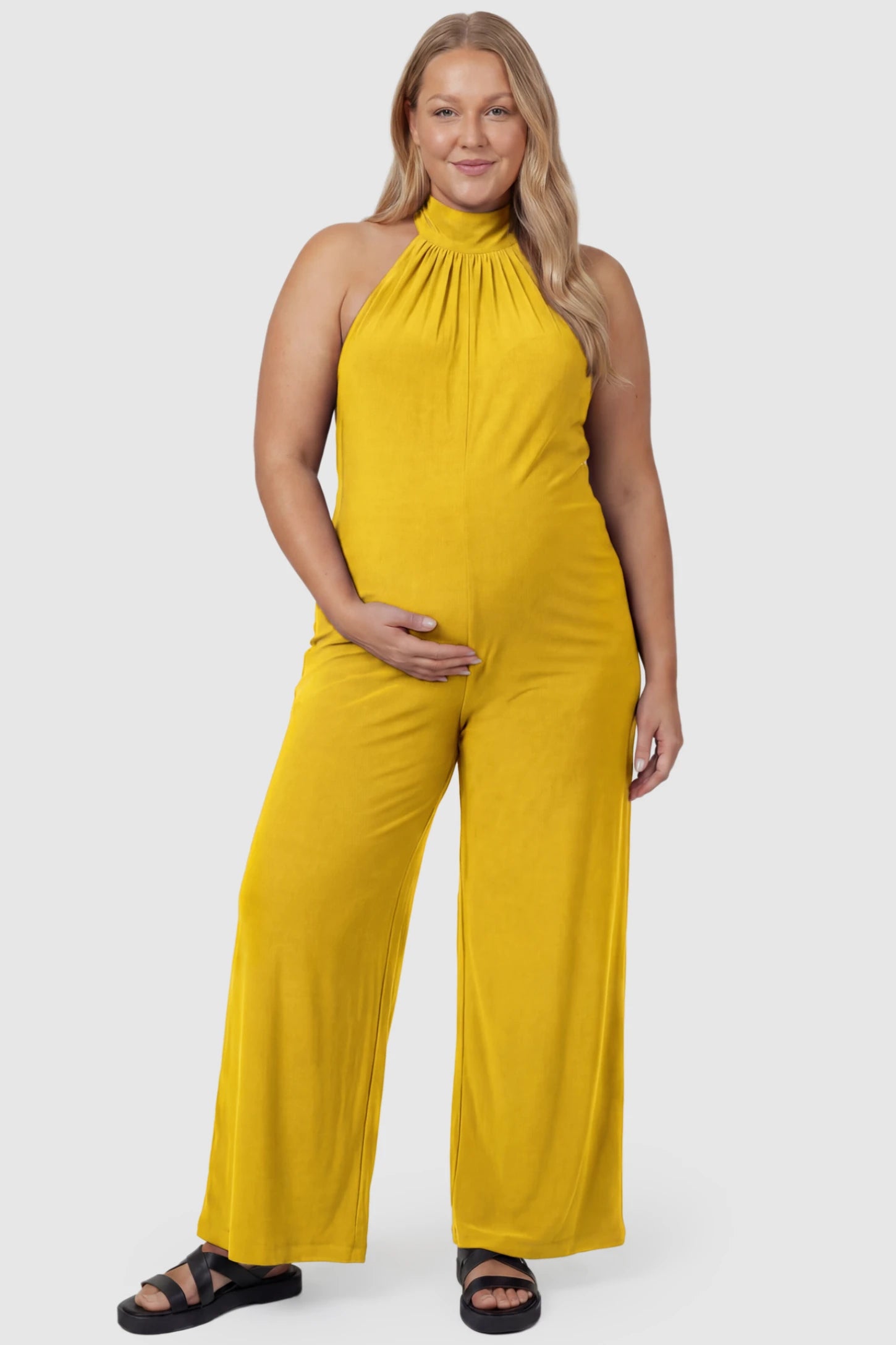 Pocketed Mock Neck Sleeveless Maternity Jumpsuit Pocketed Mock Neck Sleeveless Maternity Jumpsuit in yellow featuring a high gathered neckline, side pockets shown on a plus size pregnant model, front view.