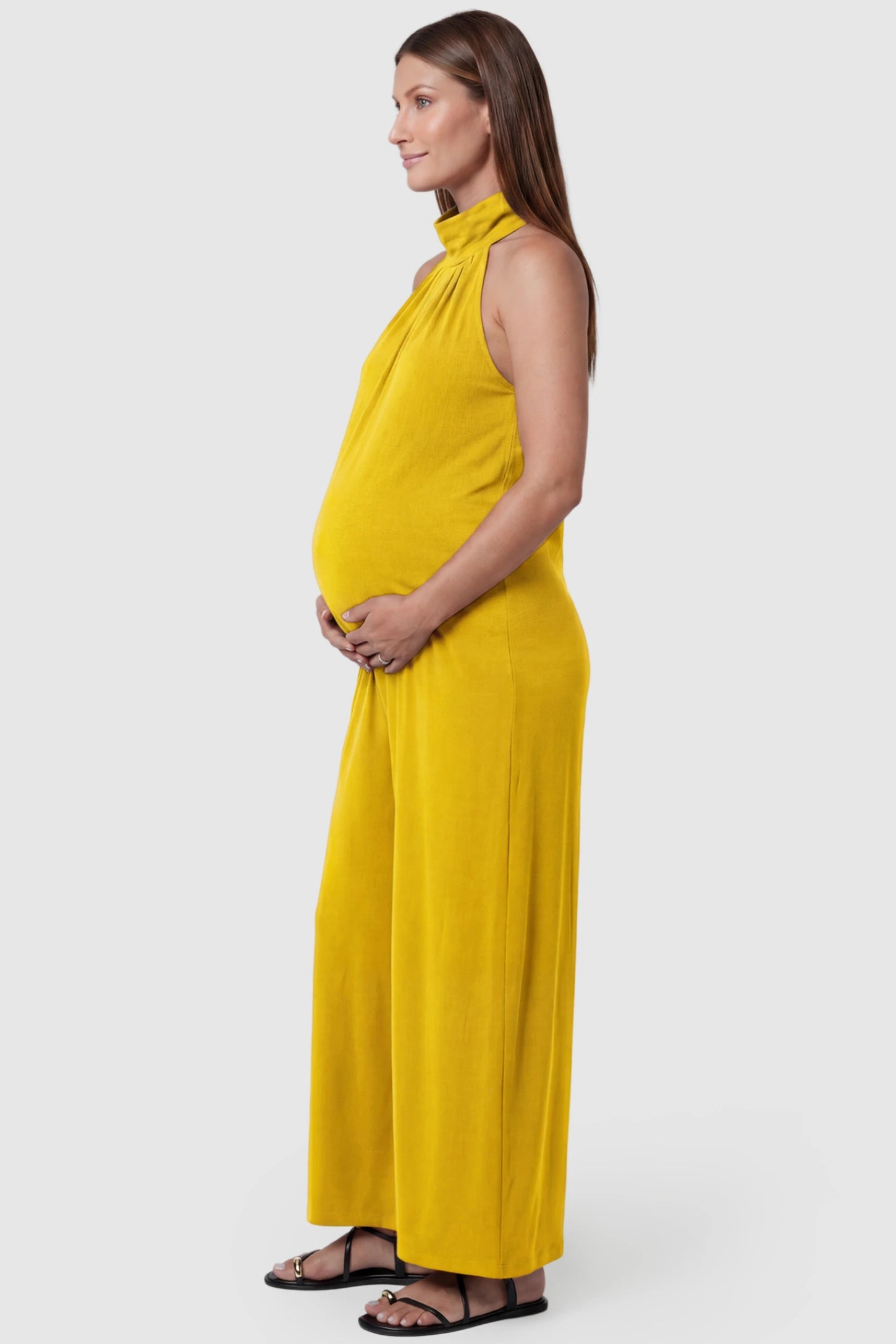 Pocketed Mock Neck Sleeveless Maternity Jumpsuit Pocketed Mock Neck Sleeveless Maternity Jumpsuit in yellow featuring a high gathered neckline, side pockets shown on a pregnant model, side view.