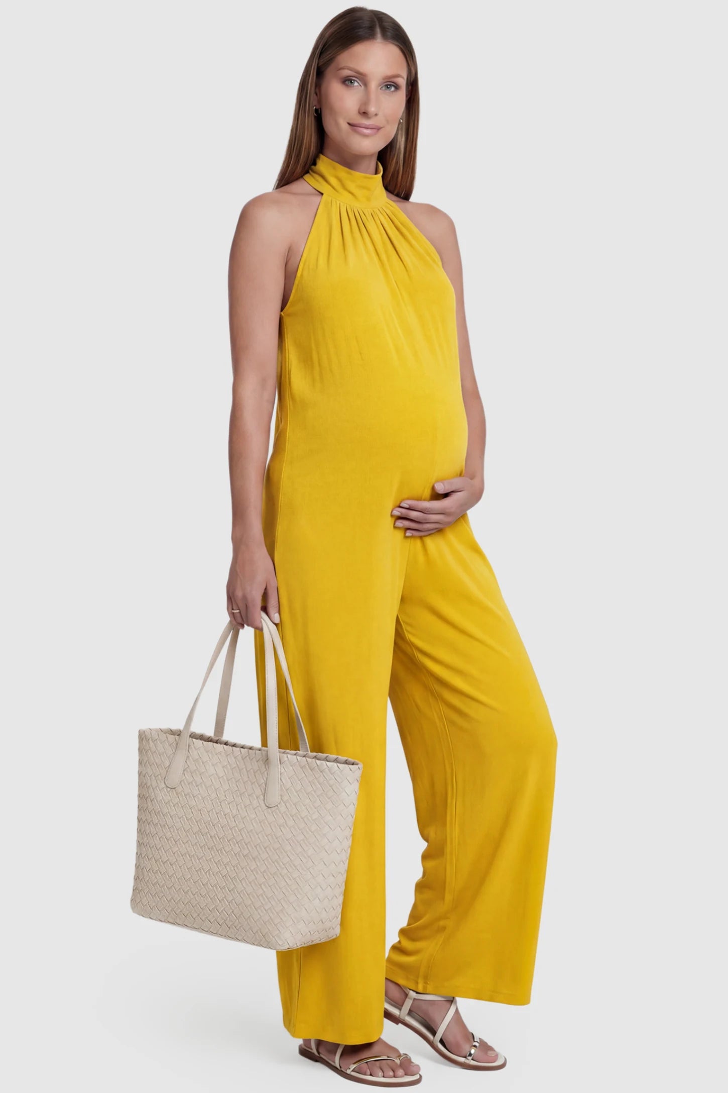 Pocketed Mock Neck Sleeveless Maternity Jumpsuit Pocketed Mock Neck Sleeveless Maternity Jumpsuit in yellow featuring a high gathered neckline, side pockets shown on a pregnant model holding a beige bag, front view.