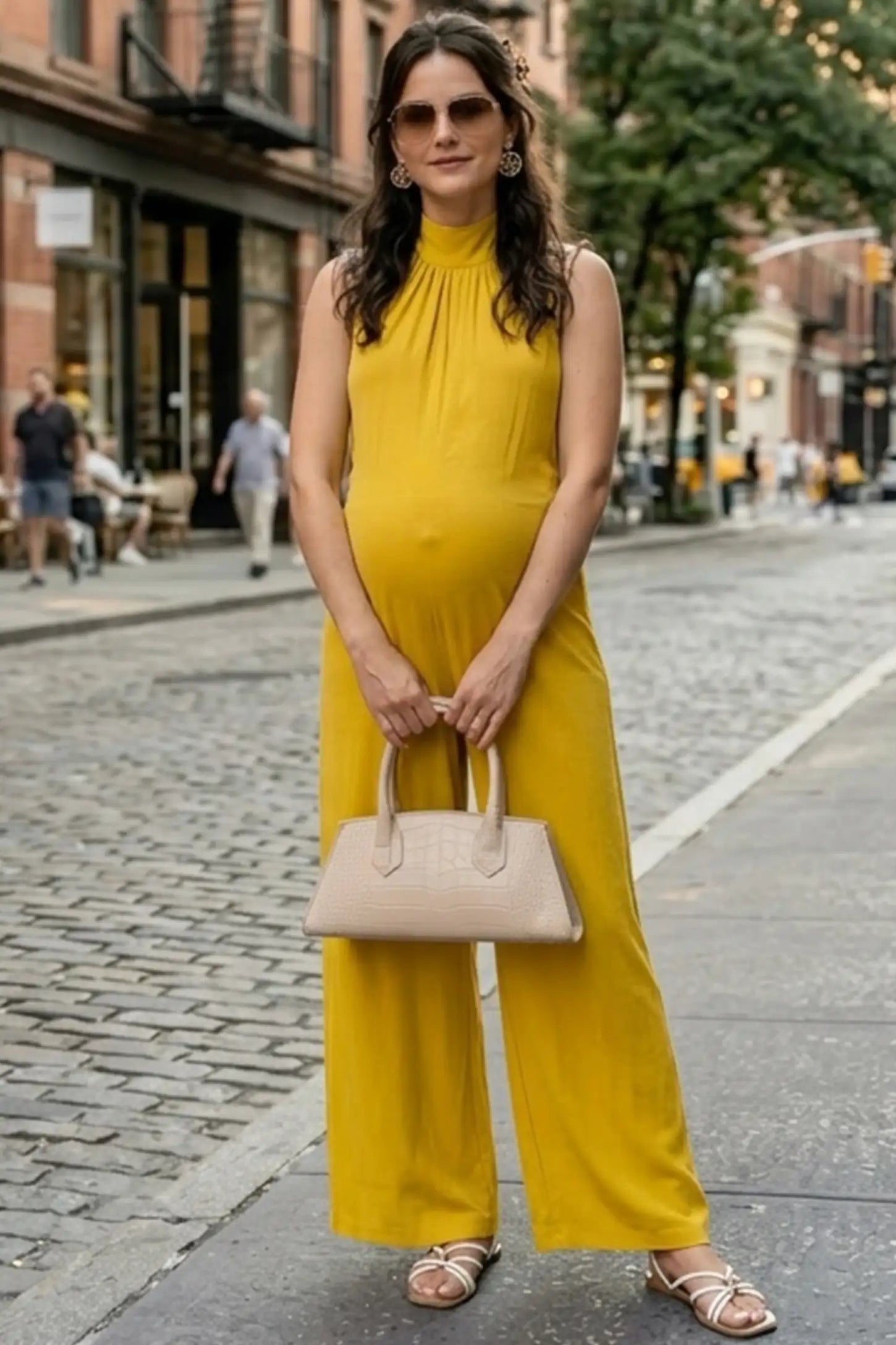Pocketed Mock Neck Sleeveless Maternity Jumpsuit Pocketed Mock Neck Sleeveless Maternity Jumpsuit in yellow featuring a high gathered neckline, side pockets shown on a pregnant woman holding a beige handbag on a city street, front view.