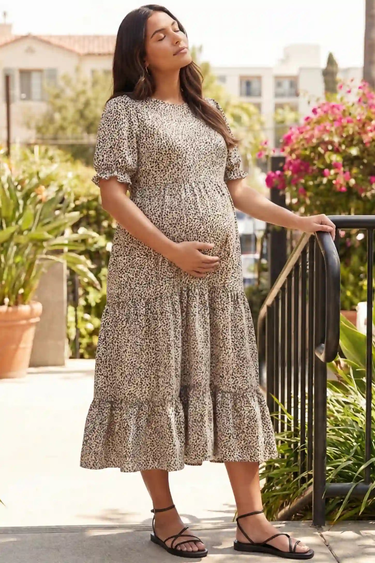 Printed Short Sleeve Tiered Maternity Midi Dress Printed Short Sleeve Tiered Maternity Midi Dress in apricot featuring round neck, tiered A-line skirt on a pregnant model cradling her belly while standing outdoors next to a black staircase railing, side view.