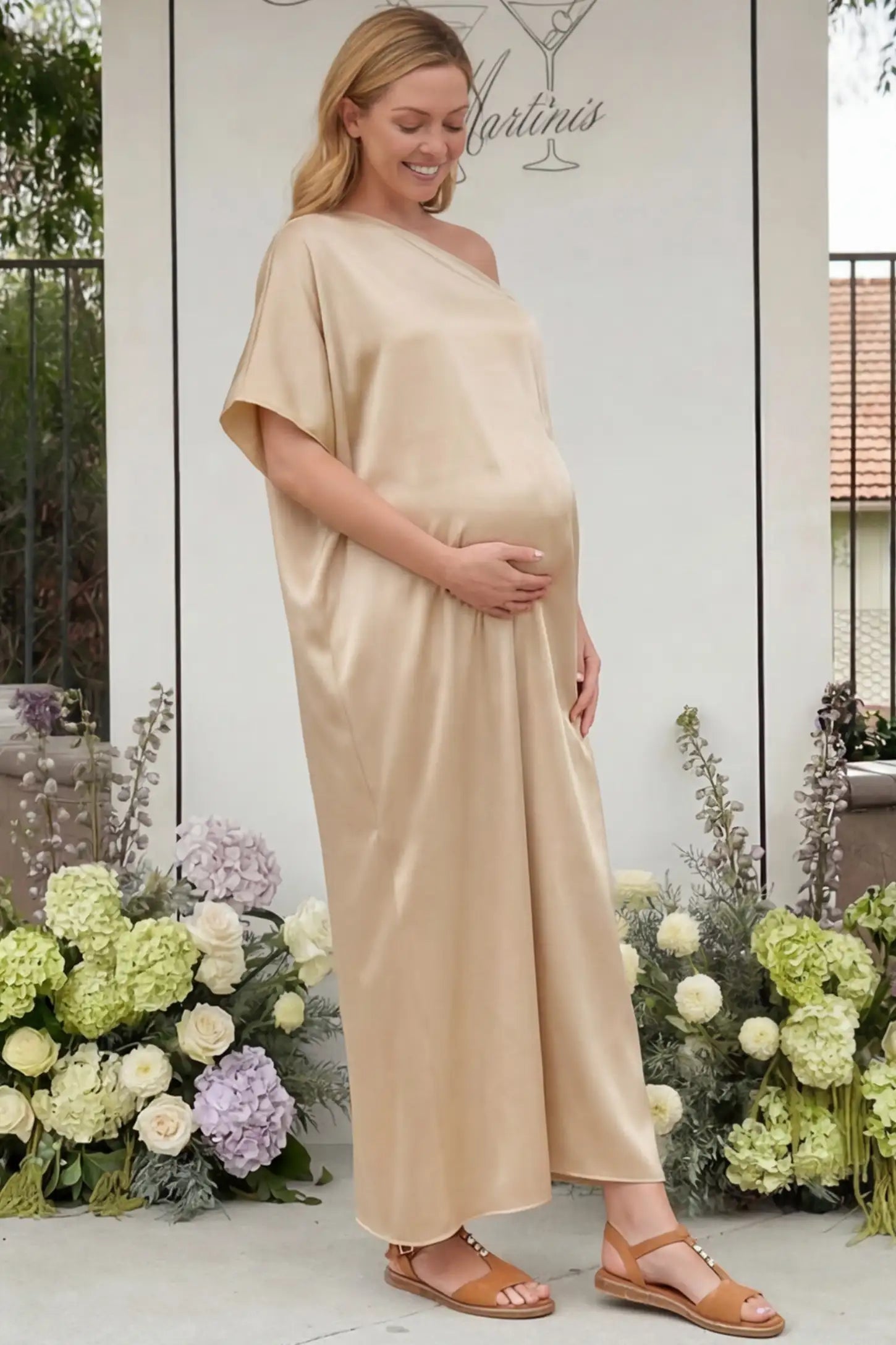 Satin One Shoulder Side Slit Maternity Maxi Dress Satin One Shoulder Side Slit Maternity Maxi Dress in Apricot featuring a draped batwing sleeve on a pregnant woman in outdoor setting, side view.