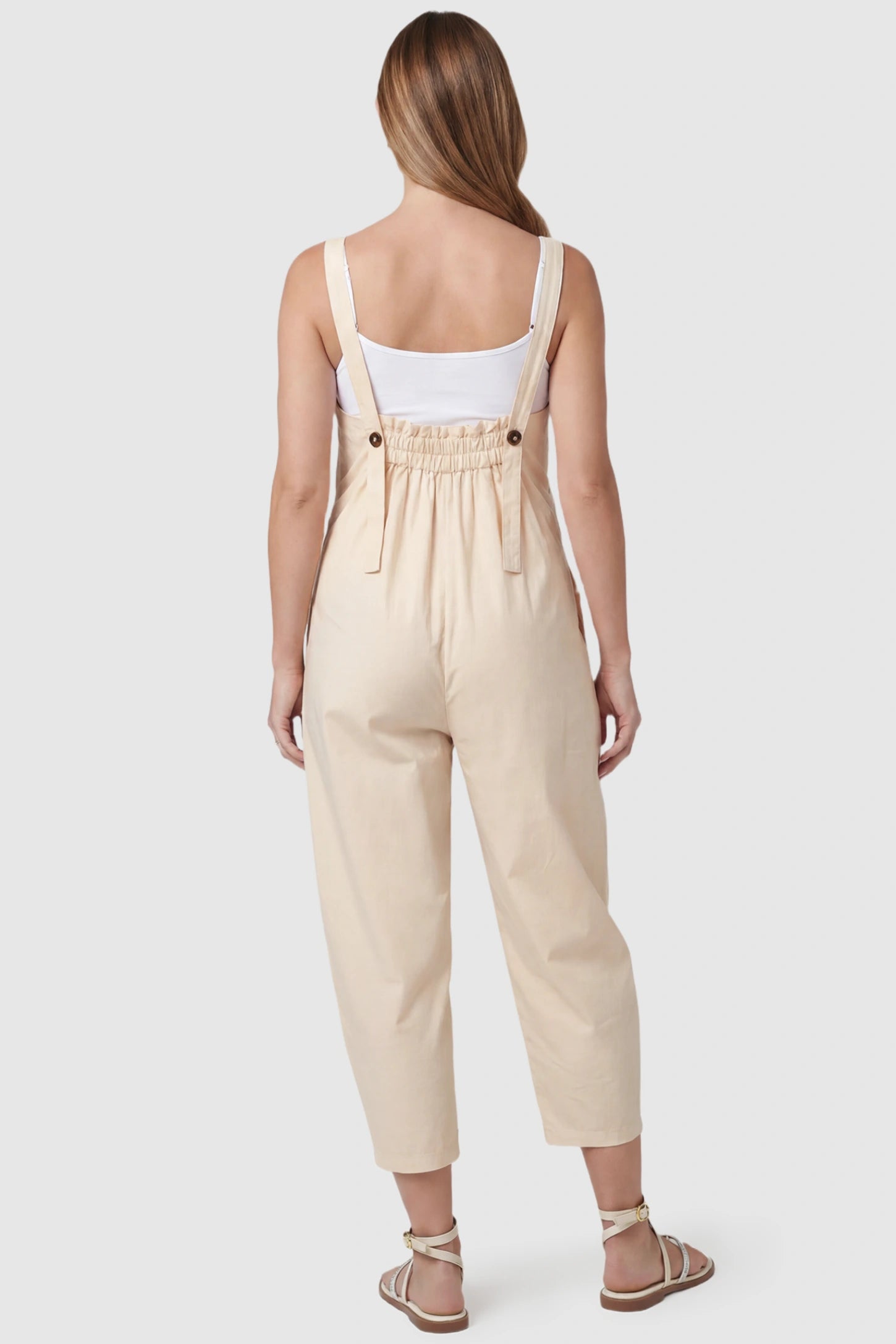 Sleeveless Balloon-Leg Maternity Jumpsuit Sleeveless Balloon-Leg Maternity Jumpsuit in Beige featuring an elasticated back and buttoned straps on a pregnant model, back view.