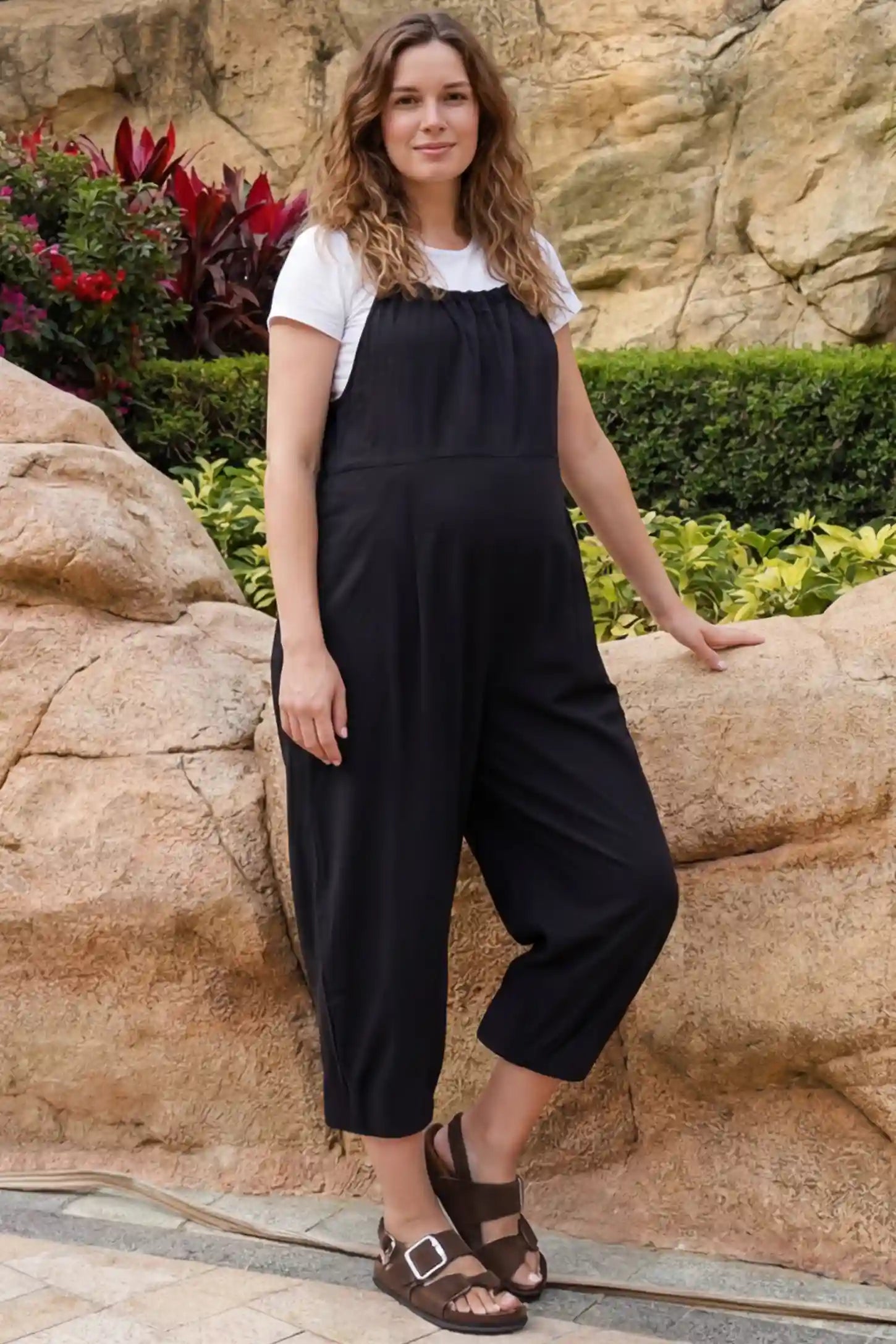 Sleeveless Balloon-Leg Maternity Jumpsuit Sleeveless Balloon-Leg Maternity Jumpsuit in Black featuring deep side pockets on a pregnant woman posing in an outdoor park setting with rocks and greenery.