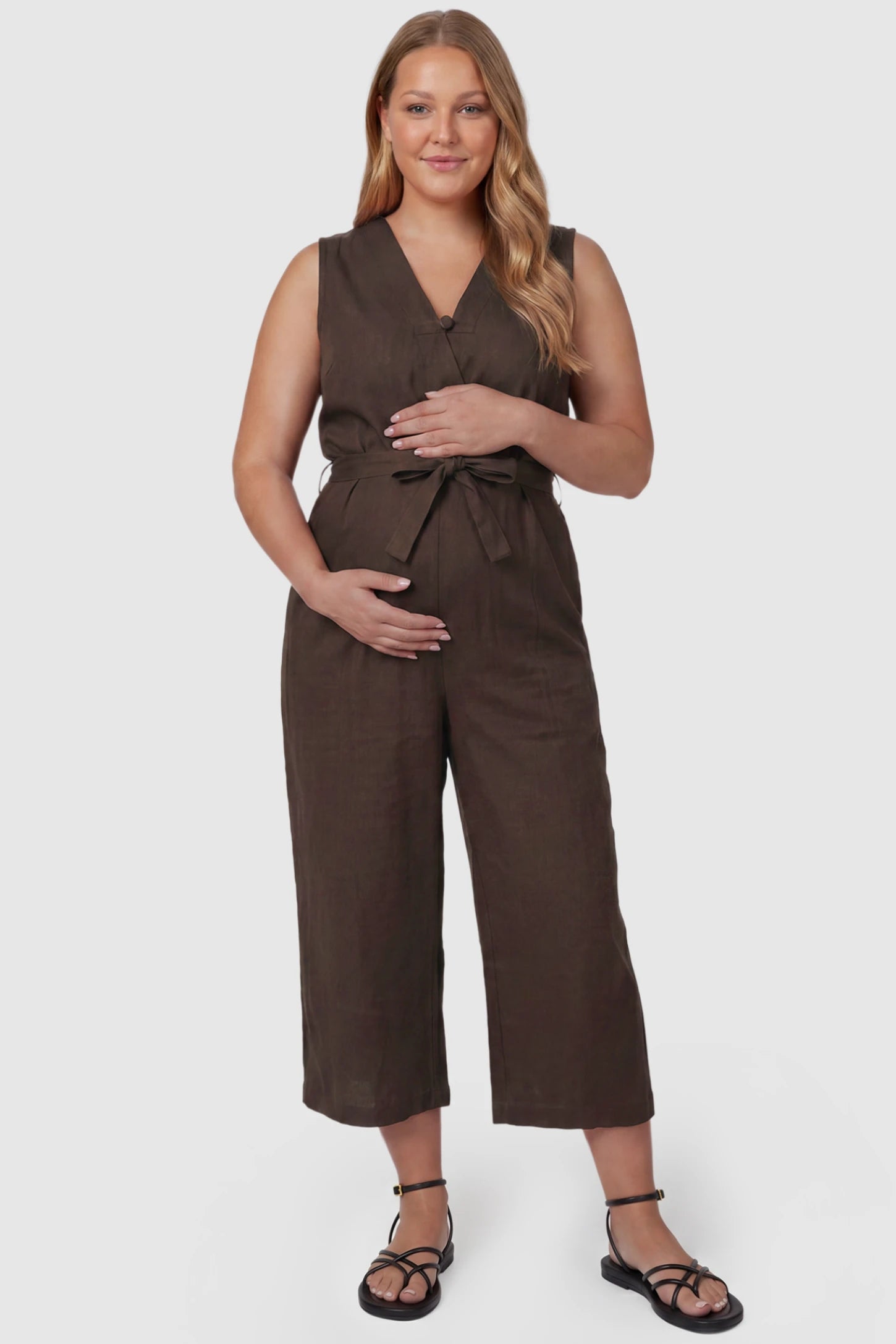 Sleeveless V-Neck Tie-Waist Maternity Jumpsuit Sleeveless V-Neck Tie-Waist Maternity Jumpsuit in Brown featuring a relaxed wide-leg design on a plus size pregnant model, front view.