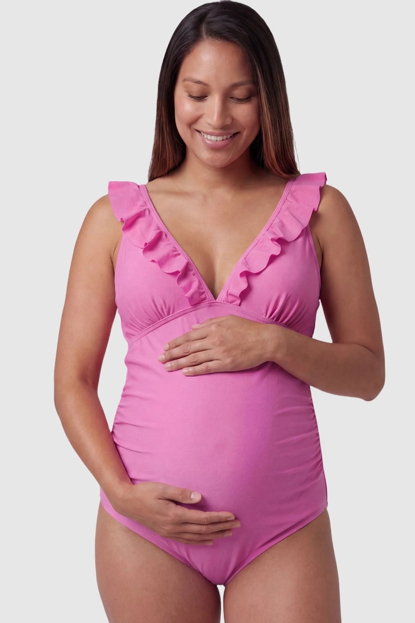Smooth Matte Ruched Maternity One-Piece Swimsuit Smooth Matte Ruched Maternity One-Piece Swimsuit in Pink featuring ruffle straps and a V-neck front on a pregnant model, front view.