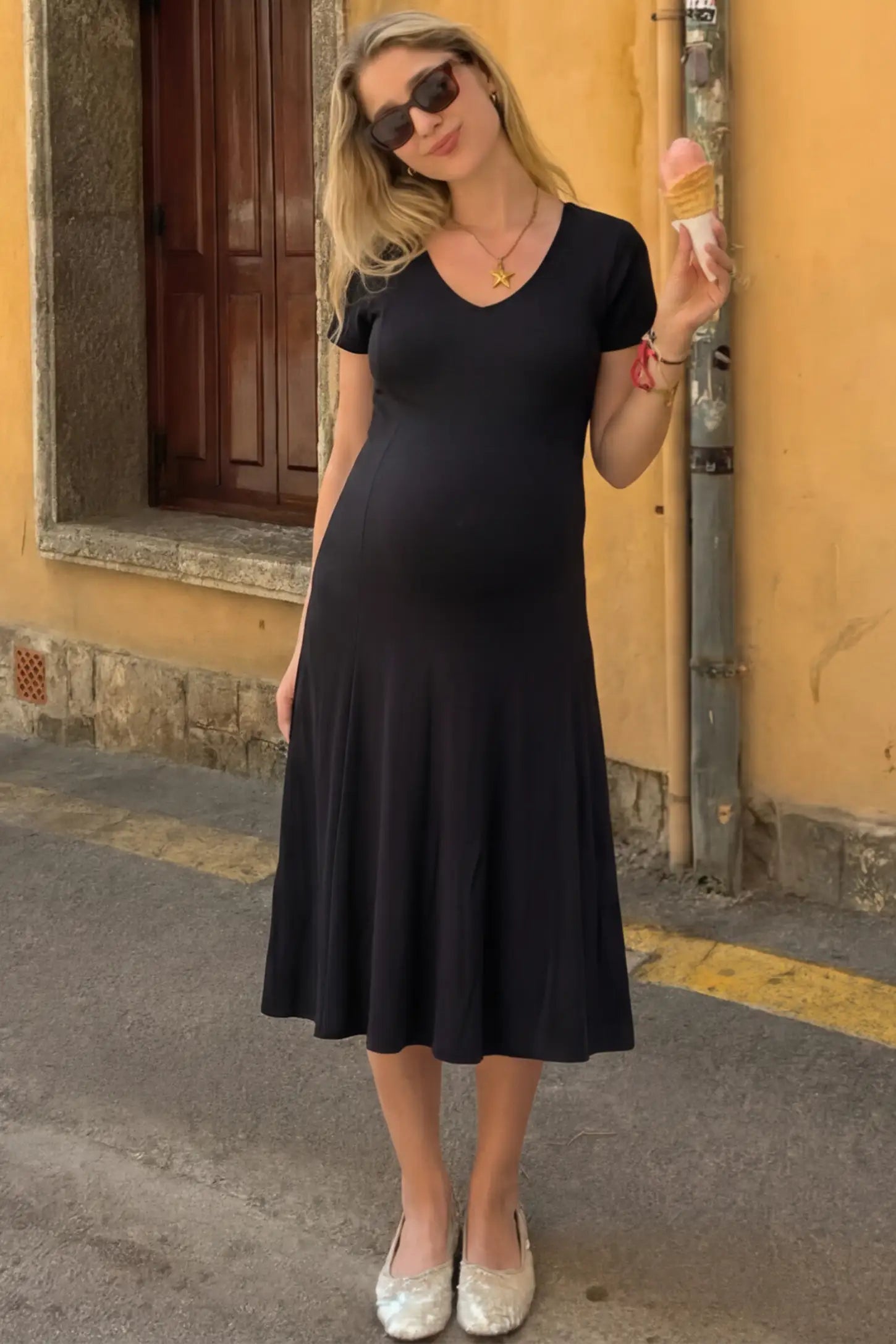 Soft Knit Stretch Short Sleeve Maternity Midi Dress Soft Knit Stretch Short Sleeve Maternity Midi Dress in Black featuring a flattering v-neck and high stretch knit on a pregnant model holding an ice cream cone outdoors, front view.