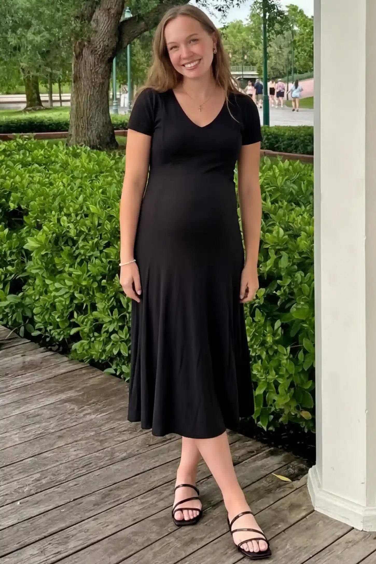 Soft Knit Stretch Short Sleeve Maternity Midi Dress Soft Knit Stretch Short Sleeve Maternity Midi Dress in Black featuring a flattering v-neck and high stretch knit on a pregnant model standing on a wooden walkway, front view.