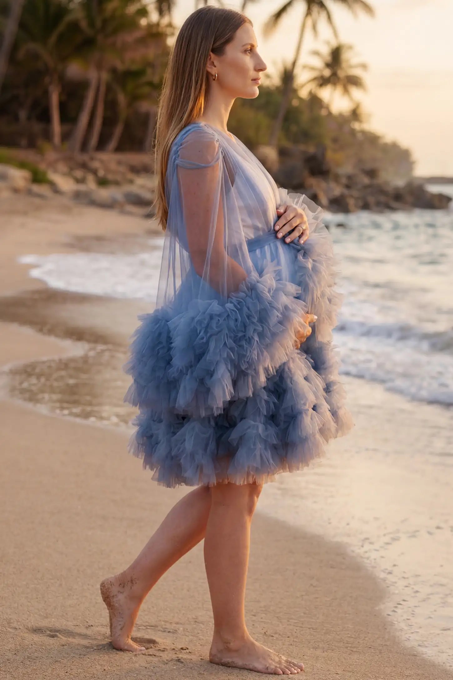 Soft Nylon Mesh Long Sleeve Maternity Photoshoot Dress Soft Nylon Mesh Long Sleeve Maternity Photoshoot Dress in Blue featuring ruffle trim layered hem on a pregnant model at the beach, side view.