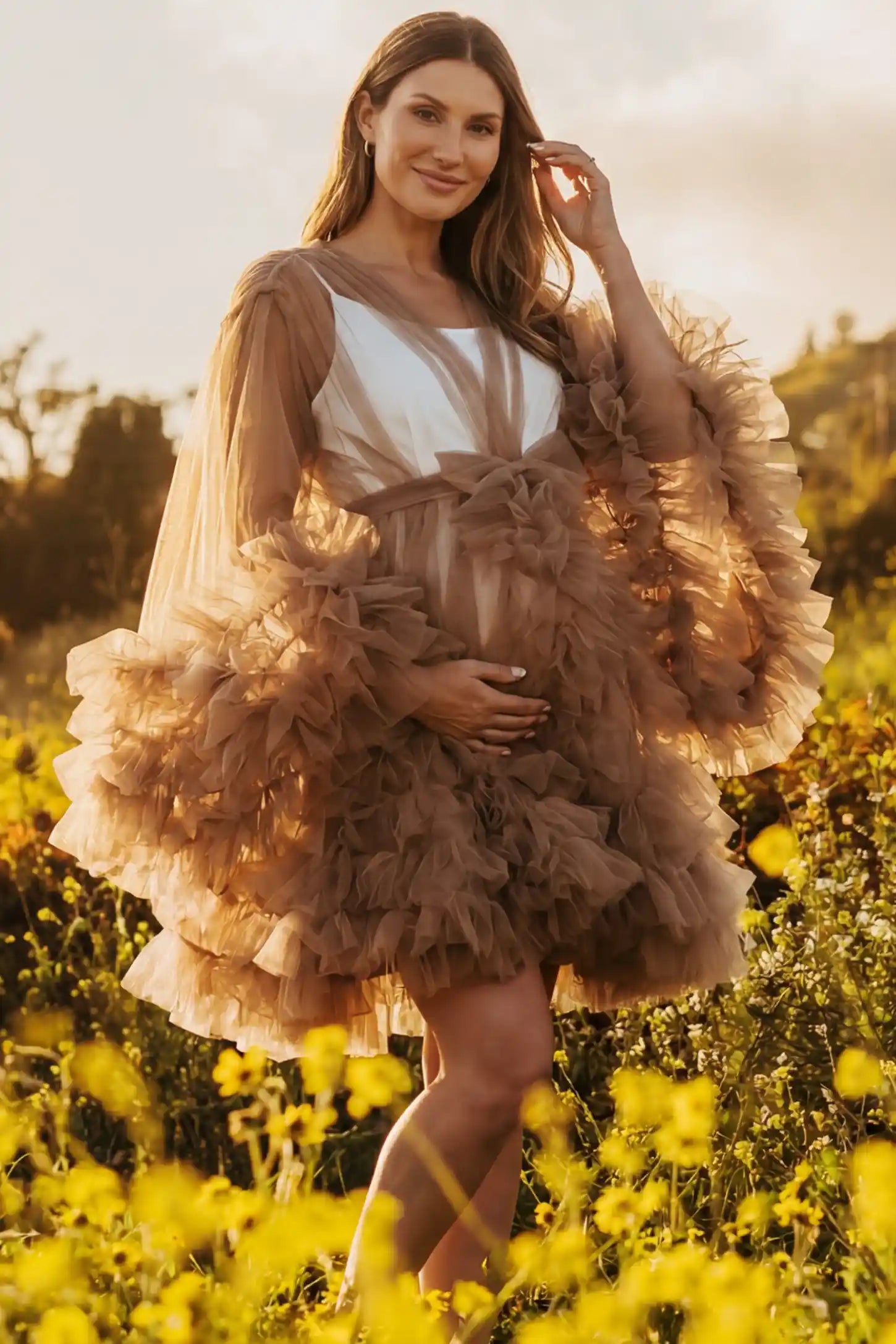Soft Nylon Mesh Long Sleeve Maternity Photoshoot Dress Soft Nylon Mesh Long Sleeve Maternity Photoshoot Dress in Camel featuring sheer V-neck, ruffle trim layered hem on a pregnant model in a wildflower field, front view.