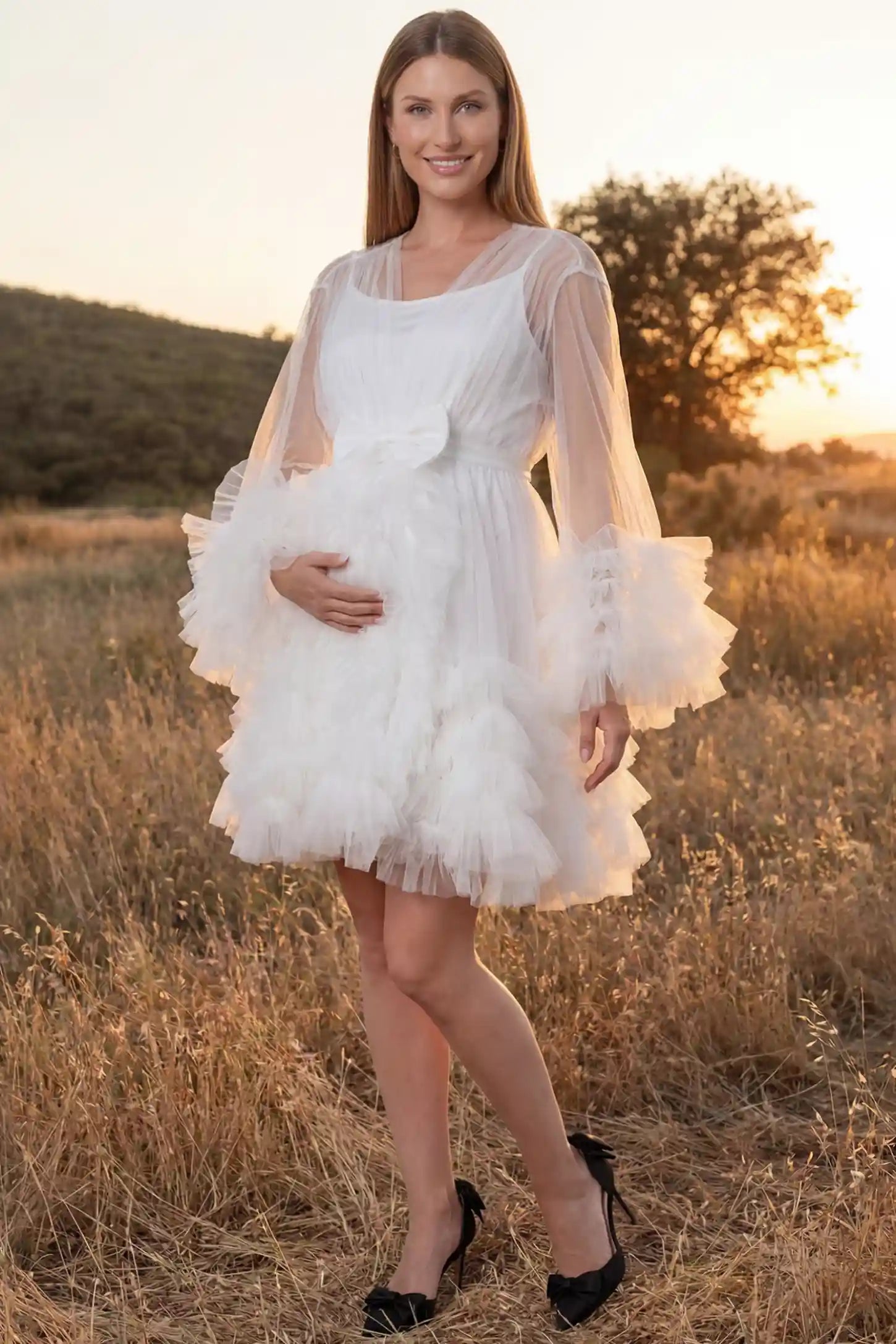 Soft Nylon Mesh Long Sleeve Maternity Photoshoot Dress Soft Nylon Mesh Long Sleeve Maternity Photoshoot Dress in White featuring sheer V-neck, ruffle trim layered hem on a pregnant model in a golden field, front view.