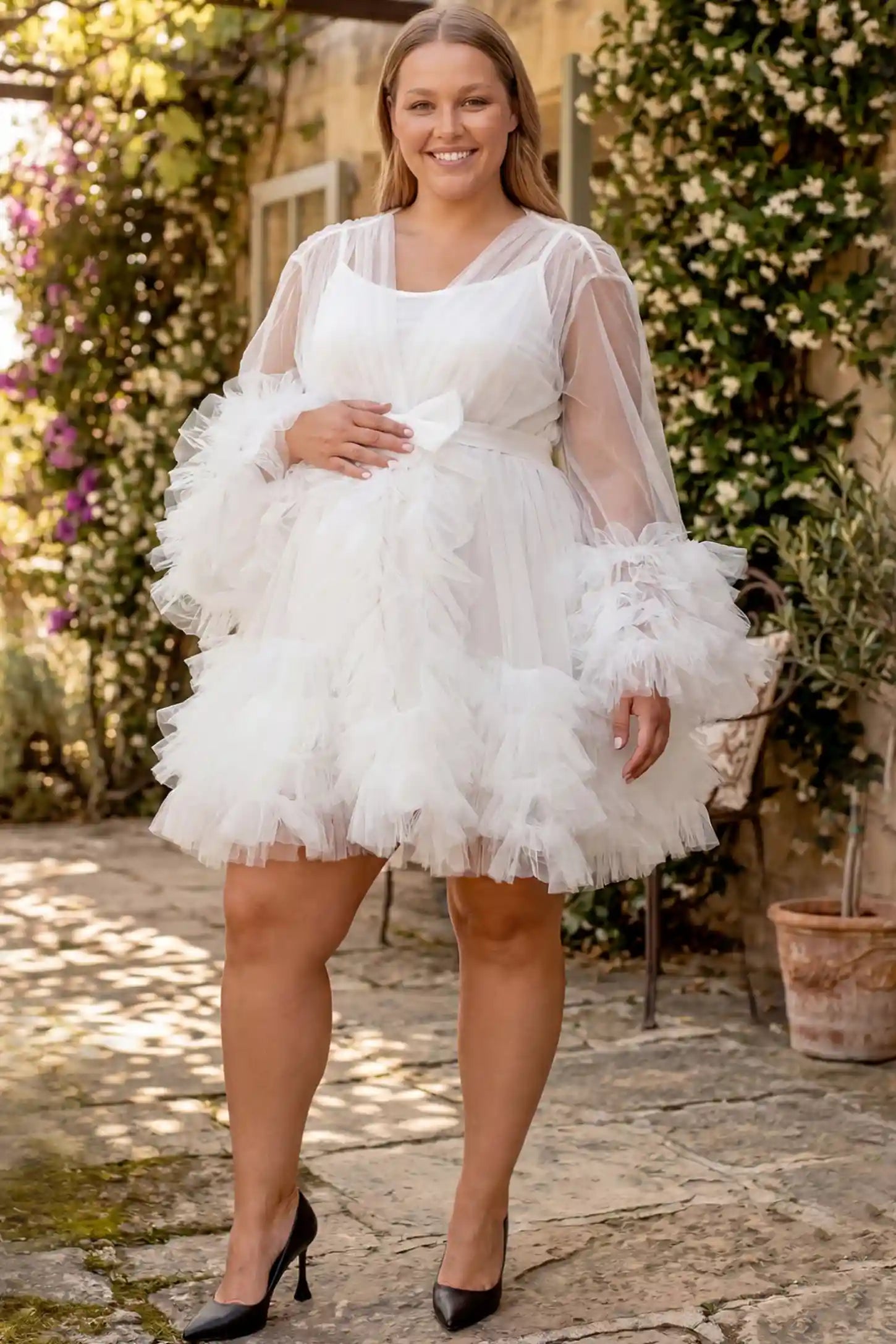 Soft Nylon Mesh Long Sleeve Maternity Photoshoot Dress Soft Nylon Mesh Long Sleeve Maternity Photoshoot Dress in White featuring sheer V-neck, ruffle trim layered hem on a plus size pregnant model in a courtyard, front view.