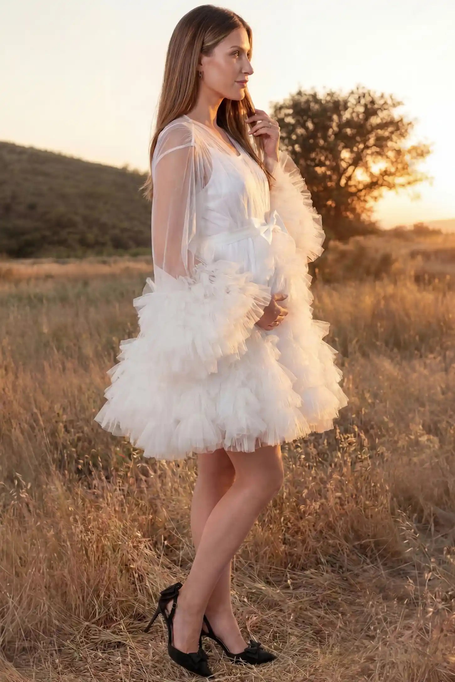 Soft Nylon Mesh Long Sleeve Maternity Photoshoot Dress Soft Nylon Mesh Long Sleeve Maternity Photoshoot Dress in White featuring ruffle trim layered hem on a pregnant model in a golden field, side view.