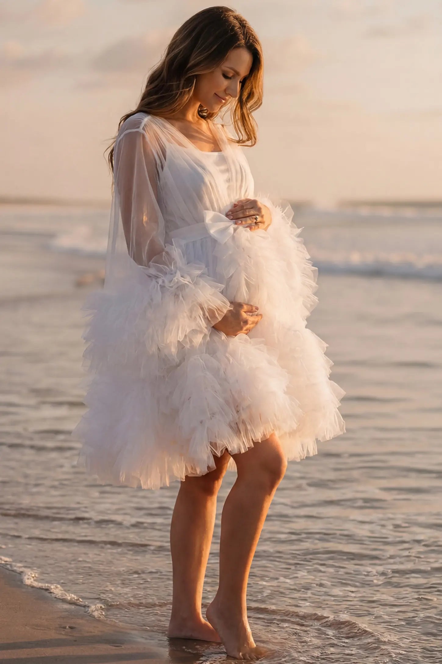 Soft Nylon Mesh Long Sleeve Maternity Photoshoot Dress Soft Nylon Mesh Long Sleeve Maternity Photoshoot Dress in White featuring sheer V-neck, ruffle trim layered hem on a pregnant model at the beach, side view.