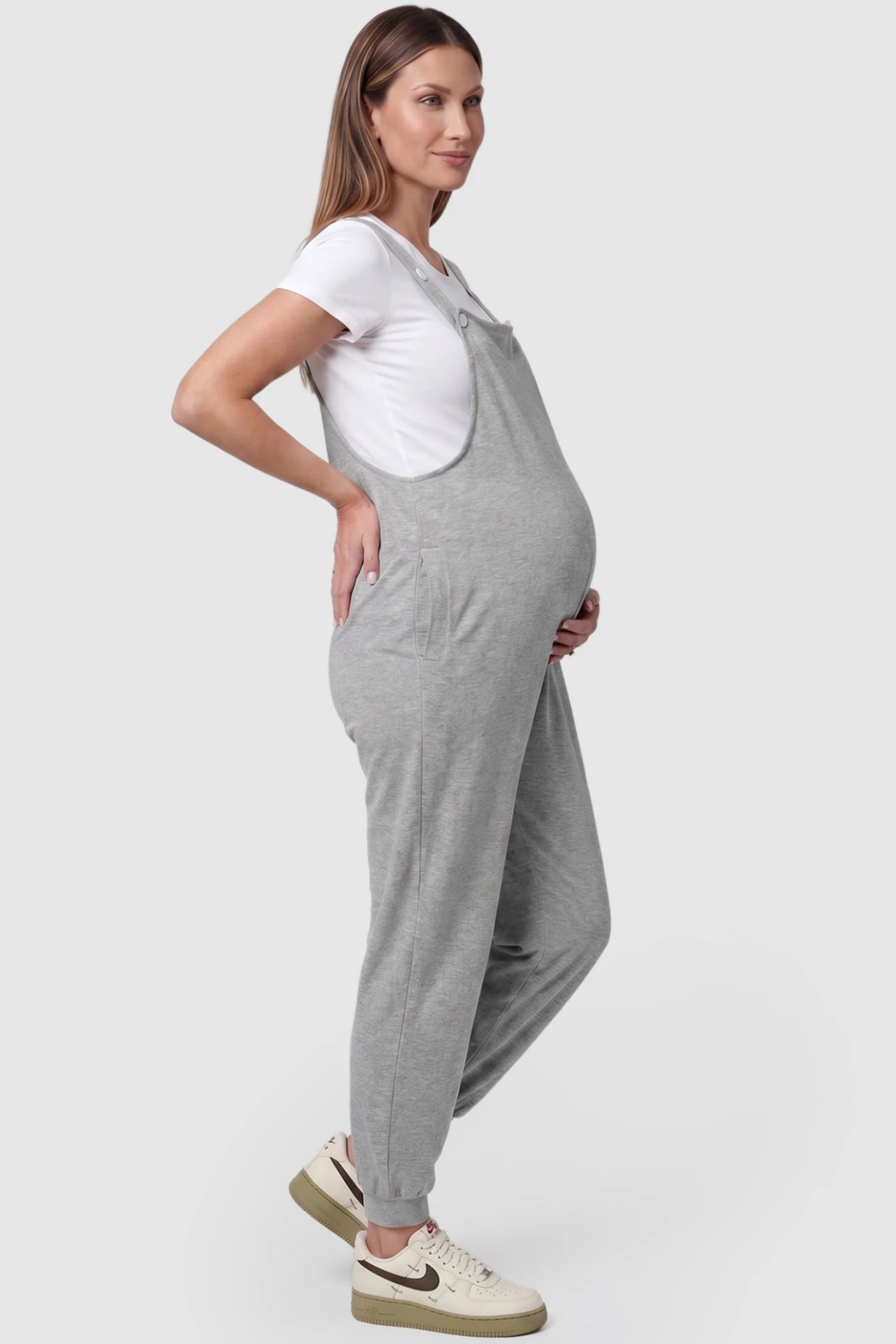 Square Neck Pocketed Maternity Jogger Jumpsuit in gray featuring button straps and side pockets on a pregnant model, side view.