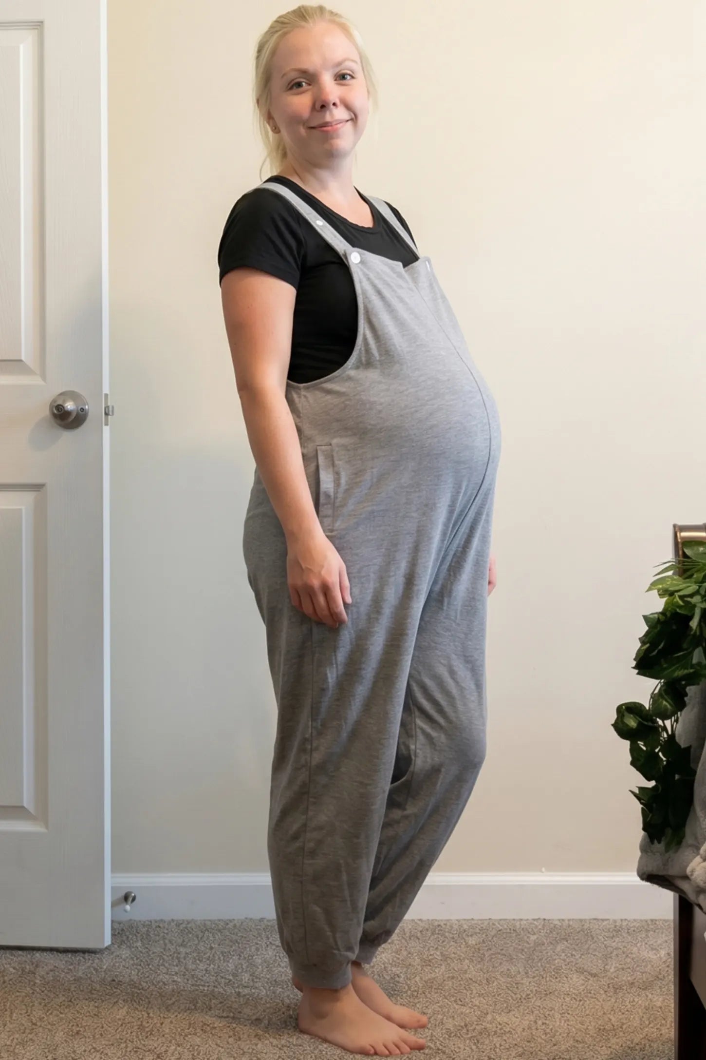 Square Neck Pocketed Maternity Jogger Jumpsuit Square Neck Pocketed Maternity Jogger Jumpsuit in gray featuring button straps and side pockets on a pregnant woman in indoor setting, side view.