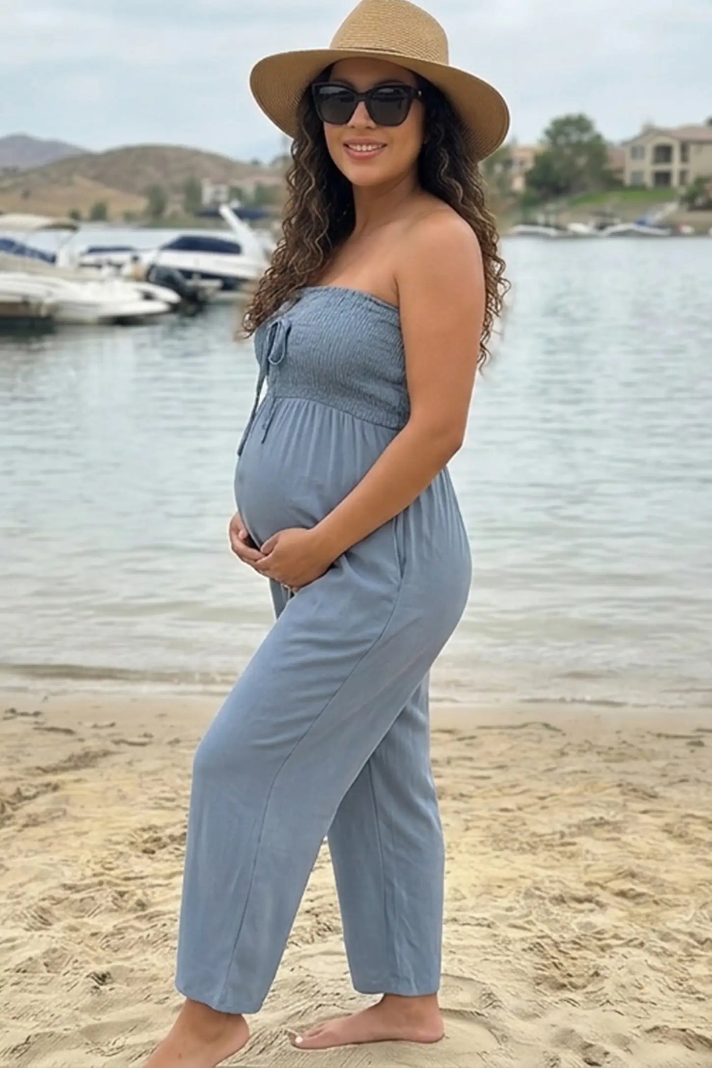 Strapless Smocked Bust Wide Leg Maternity Jumpsuit Strapless Smocked Bust Wide Leg Maternity Jumpsuit in Blue featuring a relaxed leg design on a pregnant woman in outdoor setting, side view.