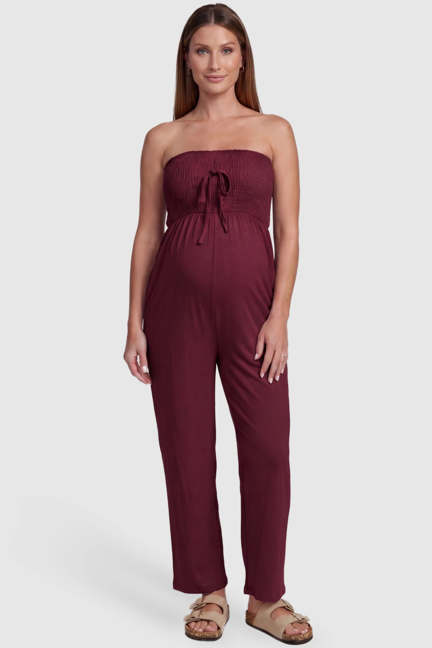 Strapless Smocked Bust Wide Leg Maternity Jumpsuit Strapless Smocked Bust Wide Leg Maternity Jumpsuit in Wine Red featuring a relaxed leg design on a pregnant model, front view.