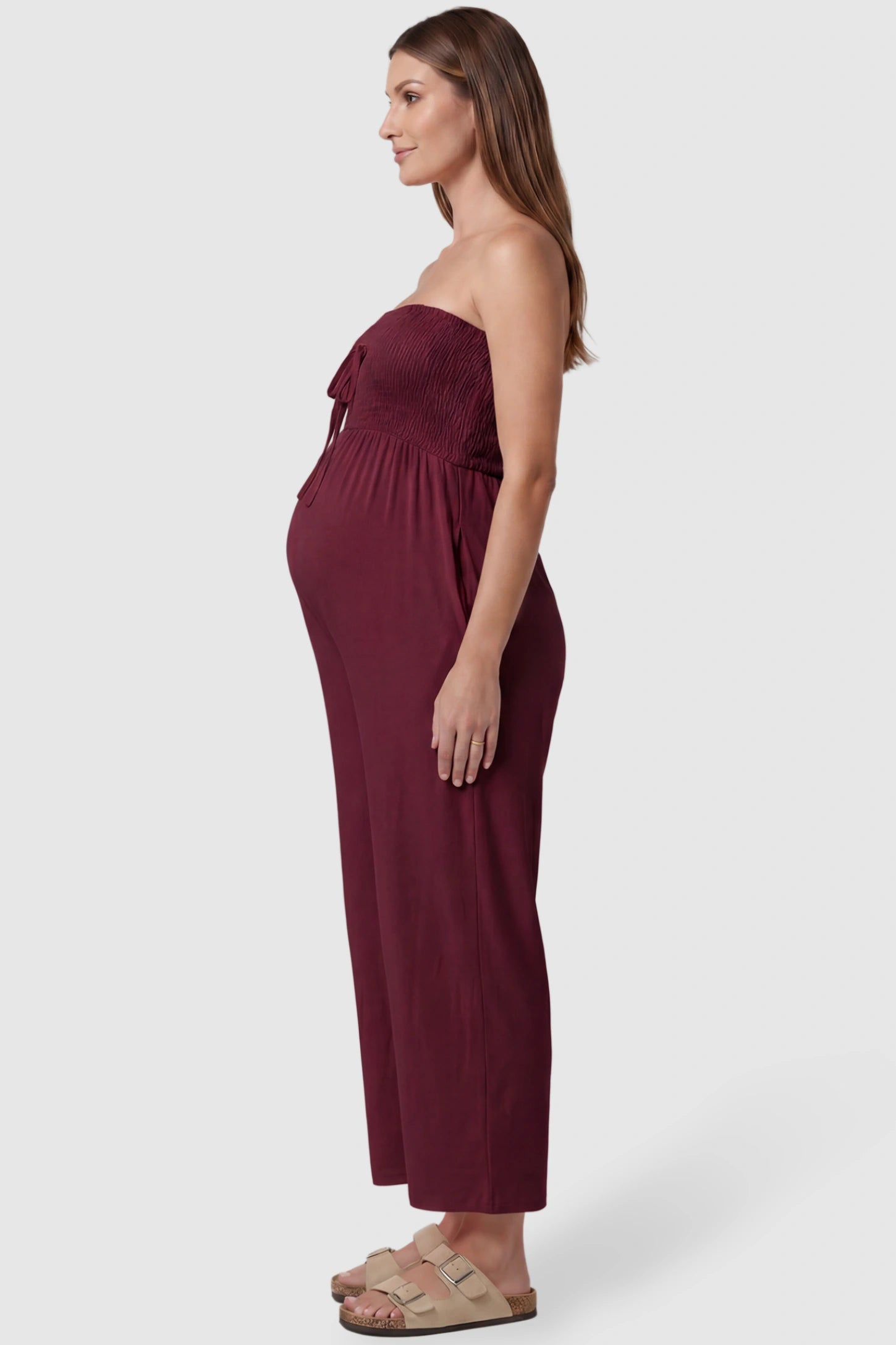 Strapless Smocked Bust Wide Leg Maternity Jumpsuit Strapless Smocked Bust Wide Leg Maternity Jumpsuit in Wine Red featuring a relaxed leg design on a pregnant model, side view.