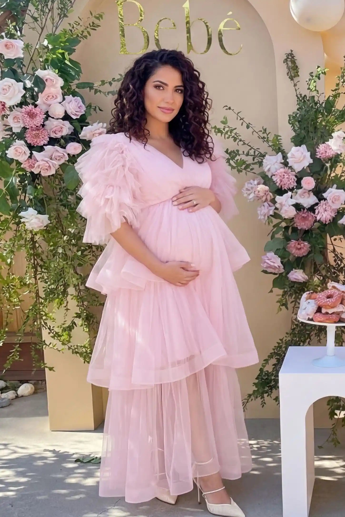 Tiered Ruffled Tulle Flutter Sleeve Maternity Maxi Gown Tiered Ruffled Tulle Flutter Sleeve Maternity Maxi Gown in Pink with sheer tulle overlay on pregnant model, garden setting, front view.