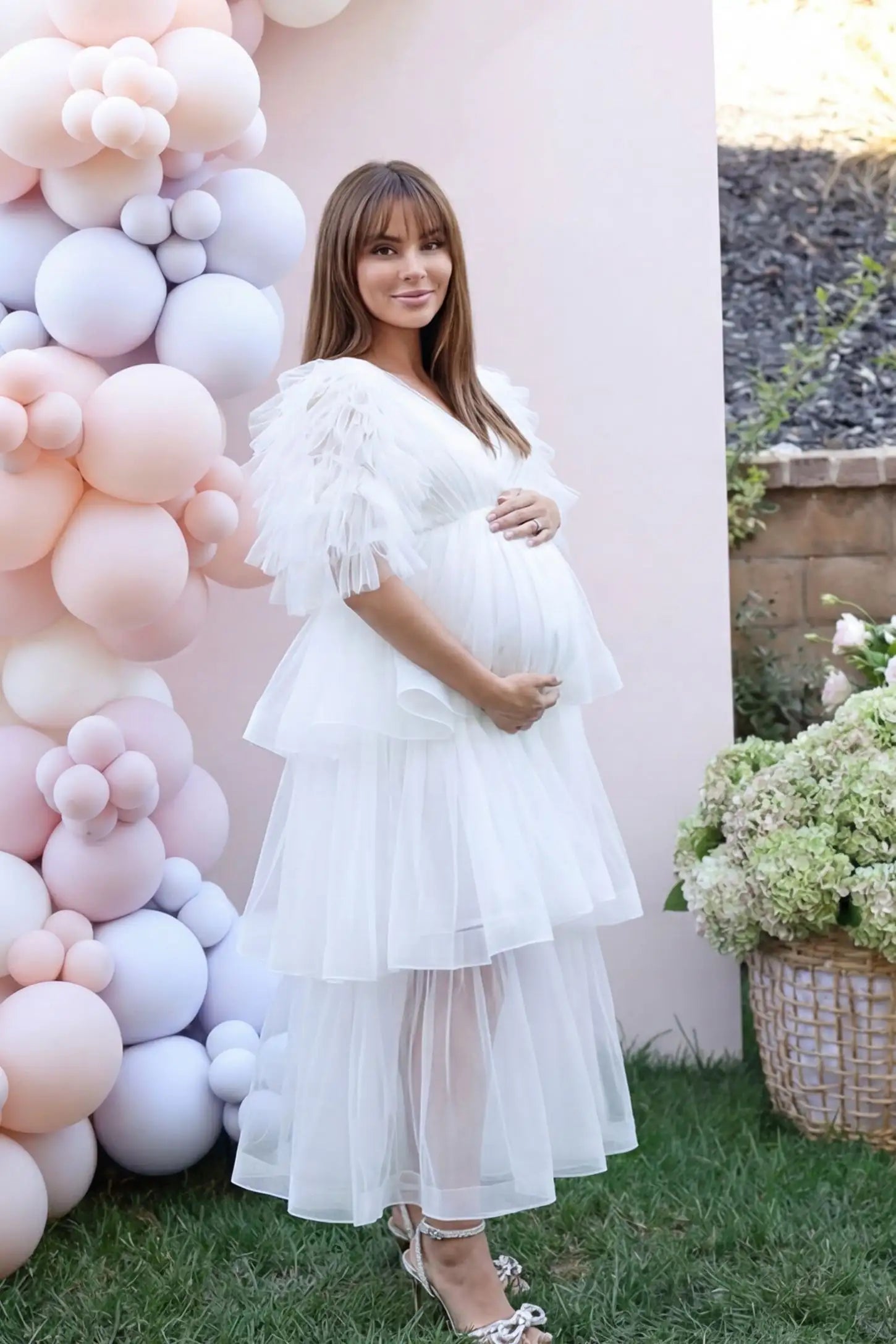 Tiered Ruffled Tulle Flutter Sleeve Maternity Maxi Gown Tiered Ruffled Tulle Flutter Sleeve Maternity Maxi Gown in White with sheer tulle overlay on pregnant model, garden setting, front view.