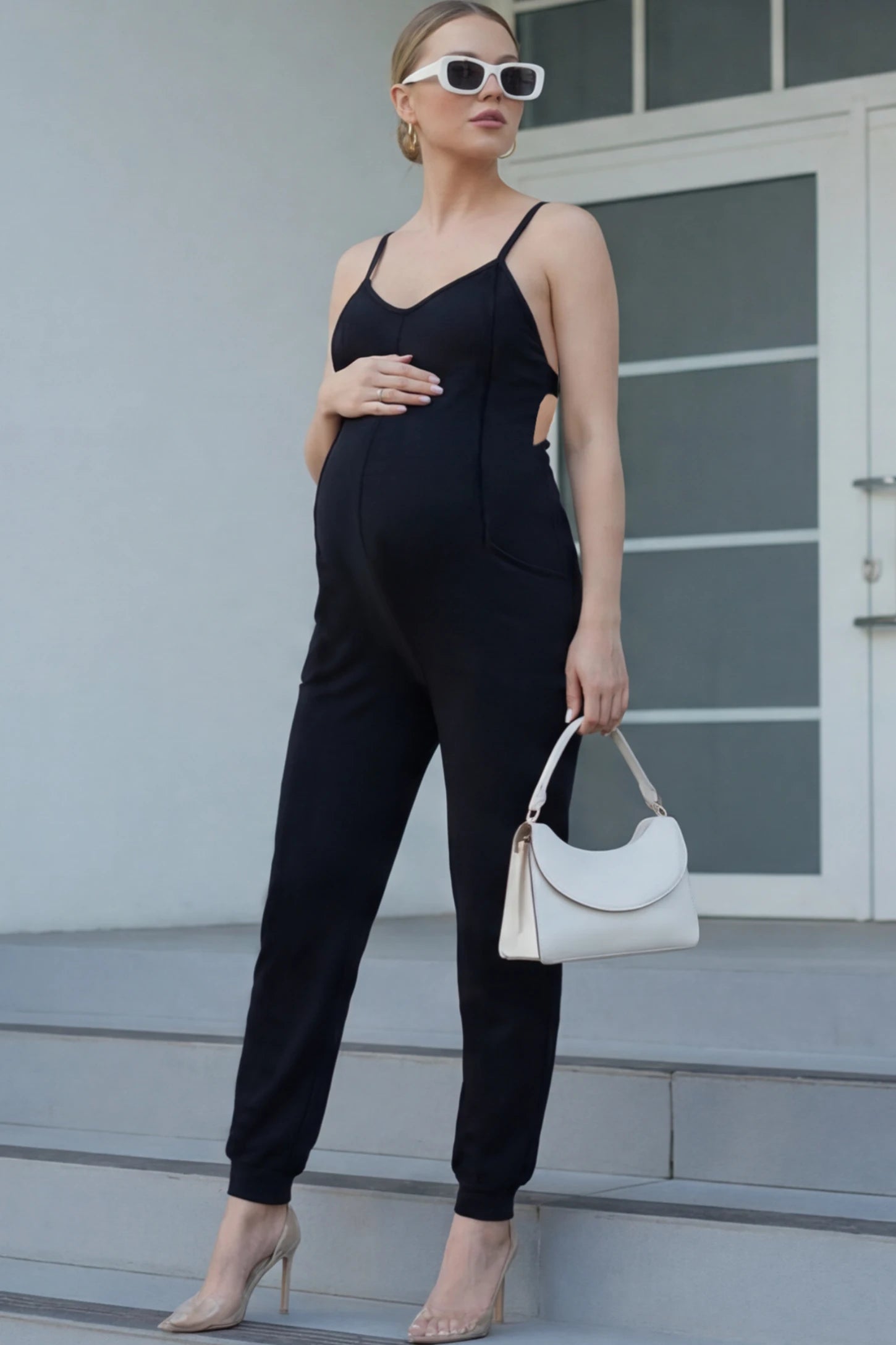 V-Neck Pocketed Maternity Jogger Jumpsuit V-Neck Pocketed Maternity Jogger Jumpsuit in Black featuring cotton-spandex jersey and side pockets on a pregnant woman in outdoor setting, front view.