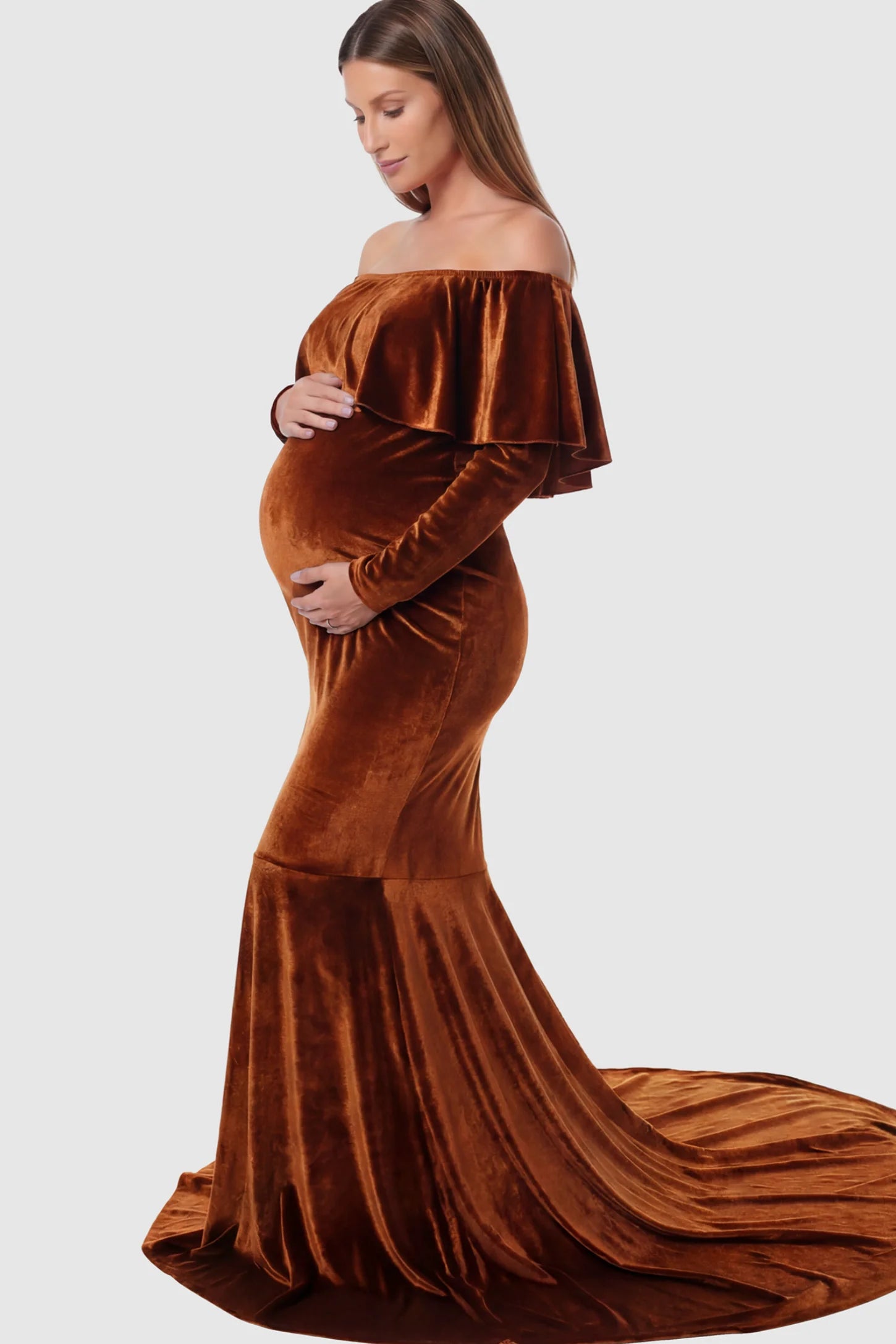 Velvet Off Shoulder Long Sleeve Maternity Mermaid Gown Velvet Off-the-Shoulder Maternity Mermaid Gown in Brown featuring a ruffled neckline and long sleeves on a pregnant model showing side.
