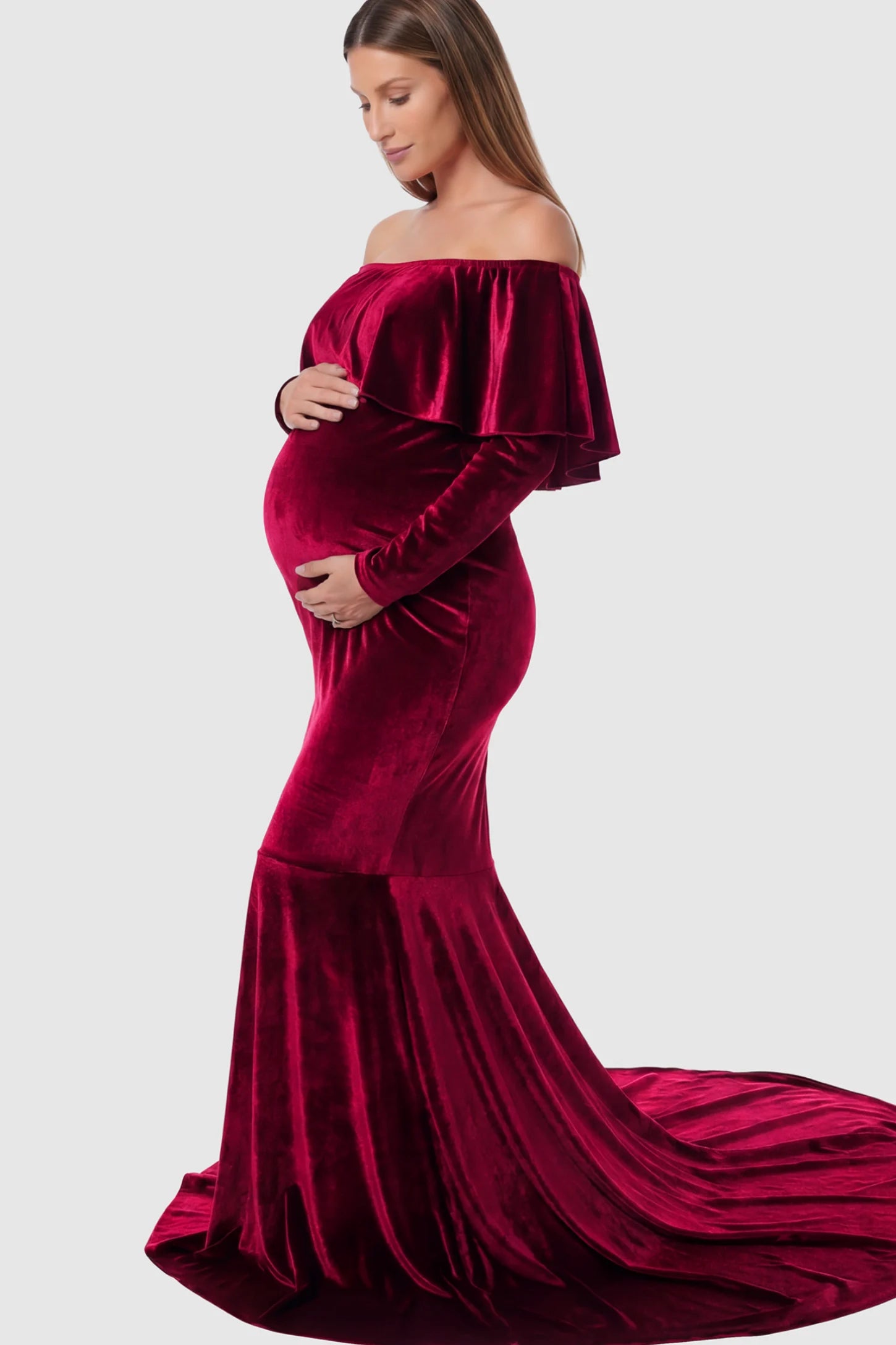 Velvet Off Shoulder Long Sleeve Maternity Mermaid Gown Velvet Off-the-Shoulder Maternity Mermaid Gown in Burgundy featuring a ruffled neckline and long sleeves on a pregnant model showing side.
